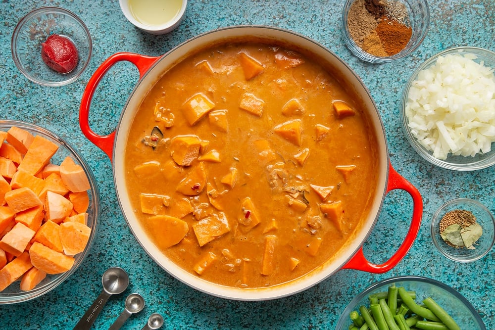 A large, shallow pan with sweet potato, coconut milk, tomato paste, fried onion, ginger, garlic, chilli and spices simmered down so the sauce is dark and thickened. The pan is surrounded by ingredients for vegan Sri Lankan curry.