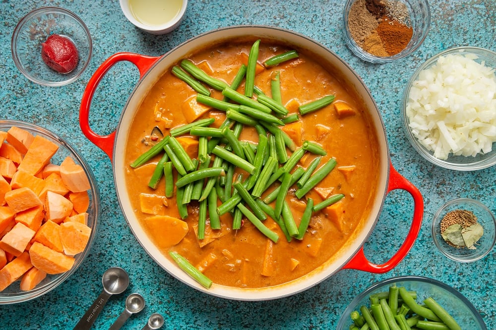 A large, shallow pan with fresh green beans added to sweet potato, coconut milk, tomato paste, fried onion, ginger, garlic, chilli and spices simmered down so the sauce is dark and thickened. The pan is surrounded by ingredients for vegan Sri Lankan curry.