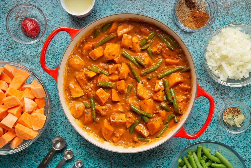 A large, shallow pan with fully cooked vegan Sri Lankan curry with sweet potato and green beans. The pan is surrounded by ingredients for vegan Sri Lankan curry.