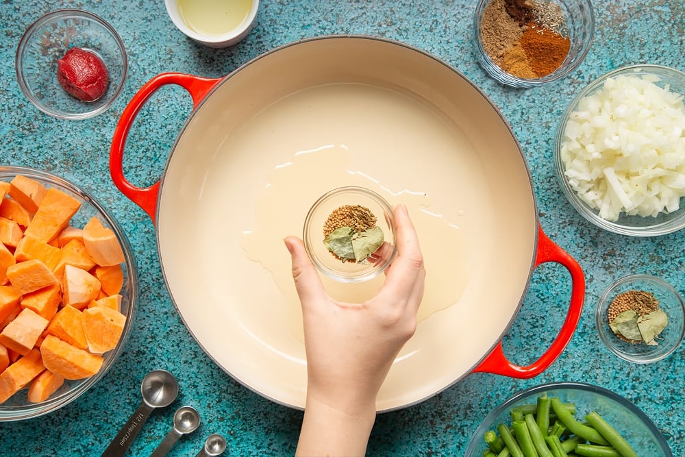 A hand holds a small bowl containing mustard seeds, curry leaves and fennel seeds over a large, shallow pan with oil in it. The pan is surrounded by ingredients for vegan Sri Lankan curry.