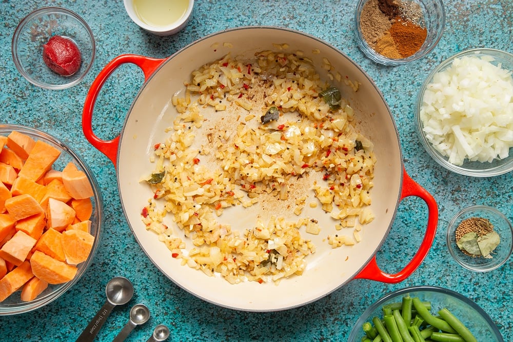 A large, shallow pan with onion, ginger, garlic, chilli and spices frying in it. The pan is surrounded by ingredients for vegan Sri Lankan curry.