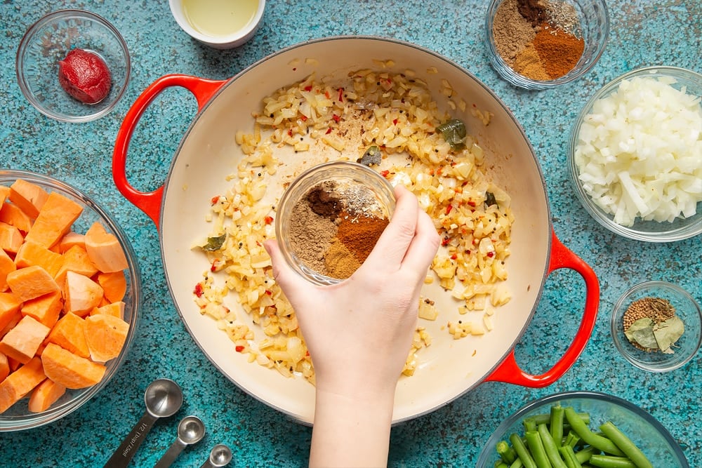 A hand holds a small bowl containing  coriander, cumin, fenugreek, cayenne, cardamom, and cloves. over a large, shallow pan with onion, ginger, garlic, chilli and spices frying in it. The pan is surrounded by ingredients for vegan Sri Lankan curry.