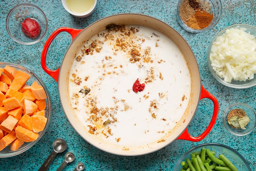 A large, shallow pan with coconut milk and tomato paste added to a mix of fried onion, ginger, garlic, chilli and spices. The pan is surrounded by ingredients for vegan Sri Lankan curry.