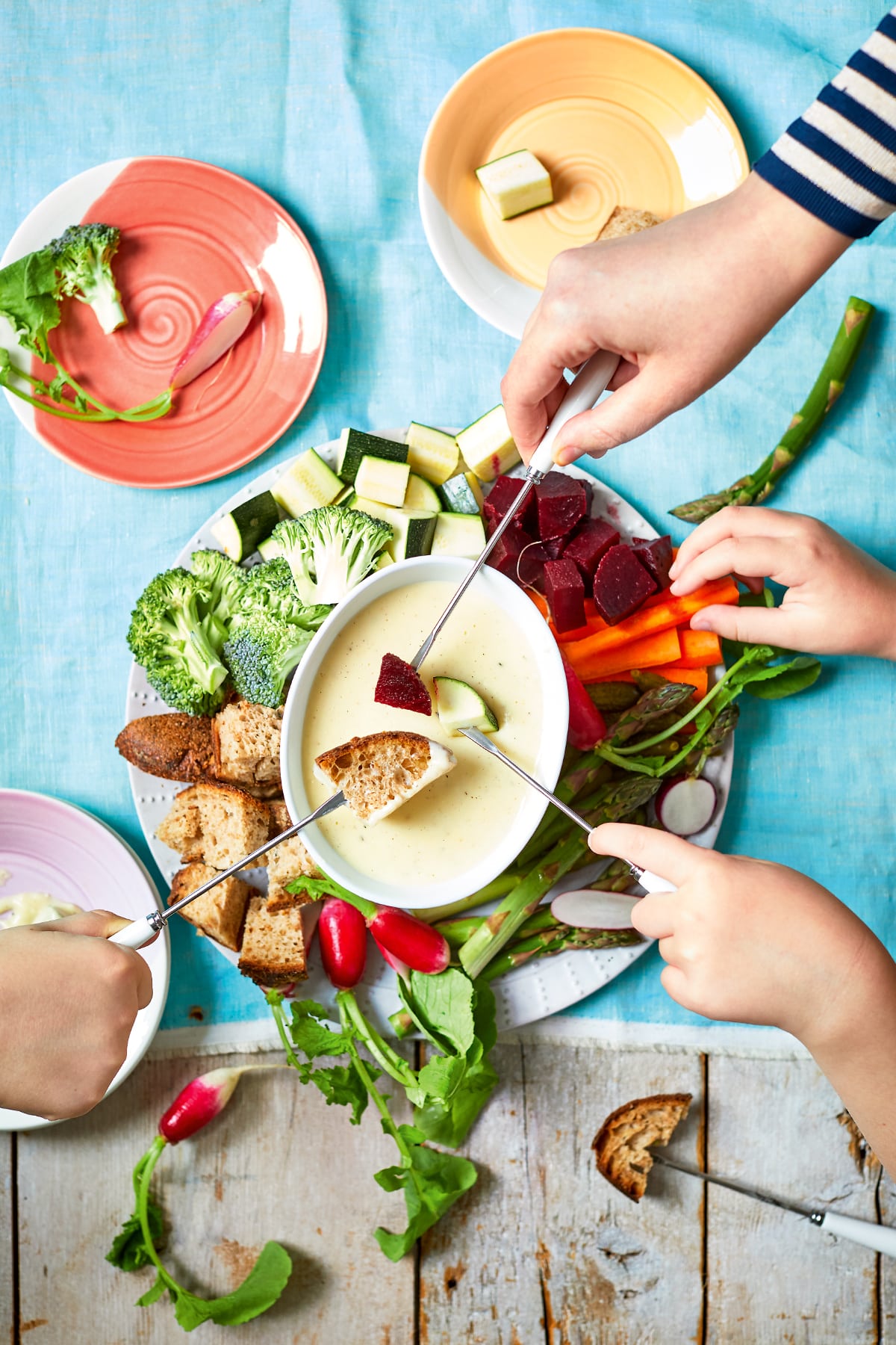 An overhead view of a vegetable fondue platter. A family of hands reaches in with fondue forks to dip bread, beetroot, courgette and more into the cheese sauce.