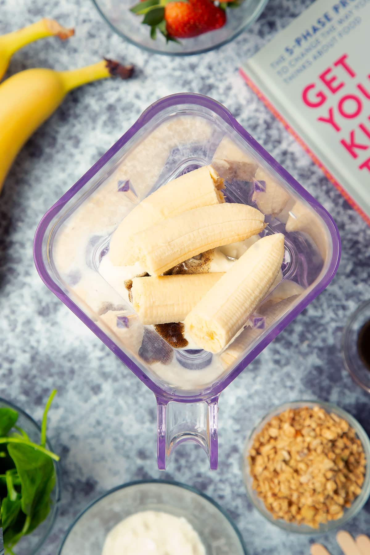 A blender is shown from above, containing yogurt, vanilla and bananas. The blender is surrounded by ingredients for fruit and veg lollies.