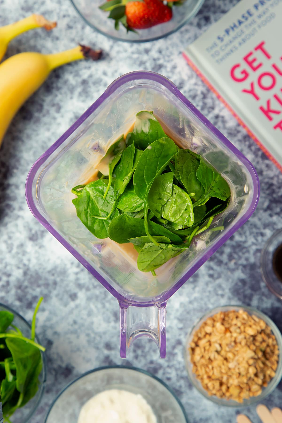 A blender containing banana yogurt mix topped with spinach. The blender is surrounded by ingredients for fruit and veg lollies.