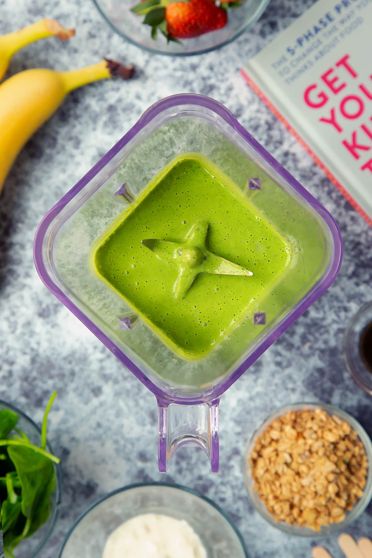 A blender containing banana yogurt spinach mix. The blender is surrounded by ingredients for fruit and veg lollies.