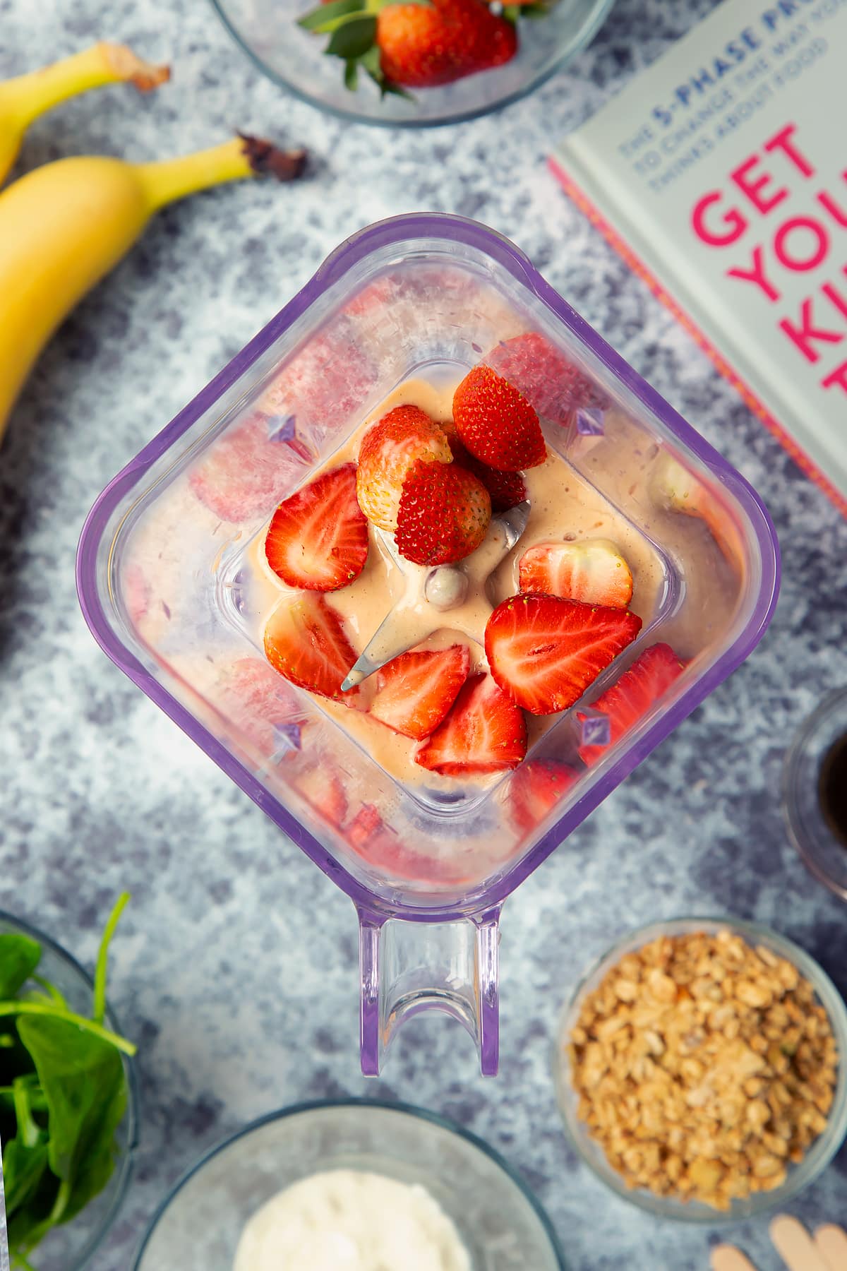 A blender containing banana yogurt mix topped with strawberries. The blender is surrounded by ingredients for fruit and veg lollies.