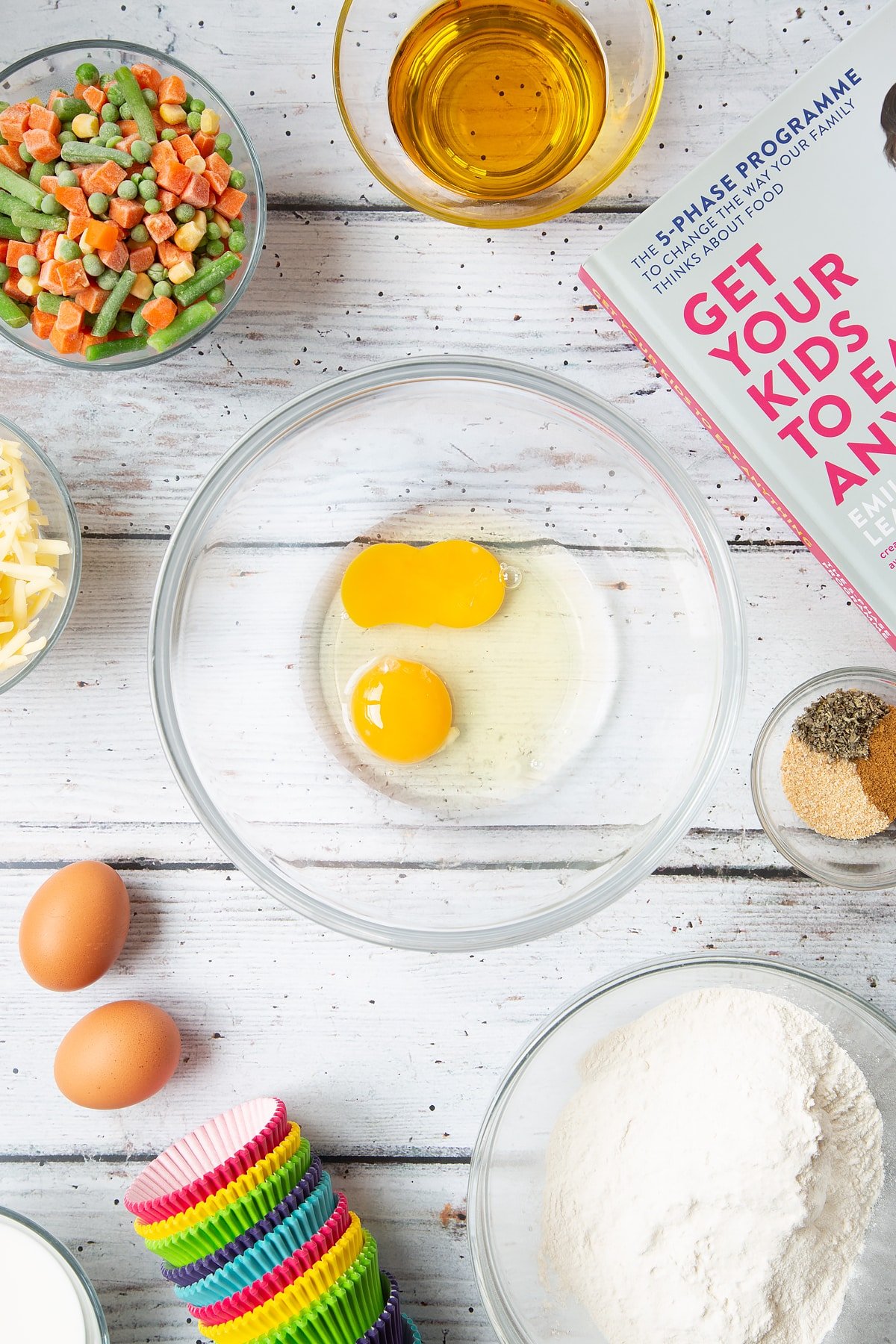 A mixing bowl containing two eggs. The bowl is surrounded by the ingredients for savoury vegetable muffins.