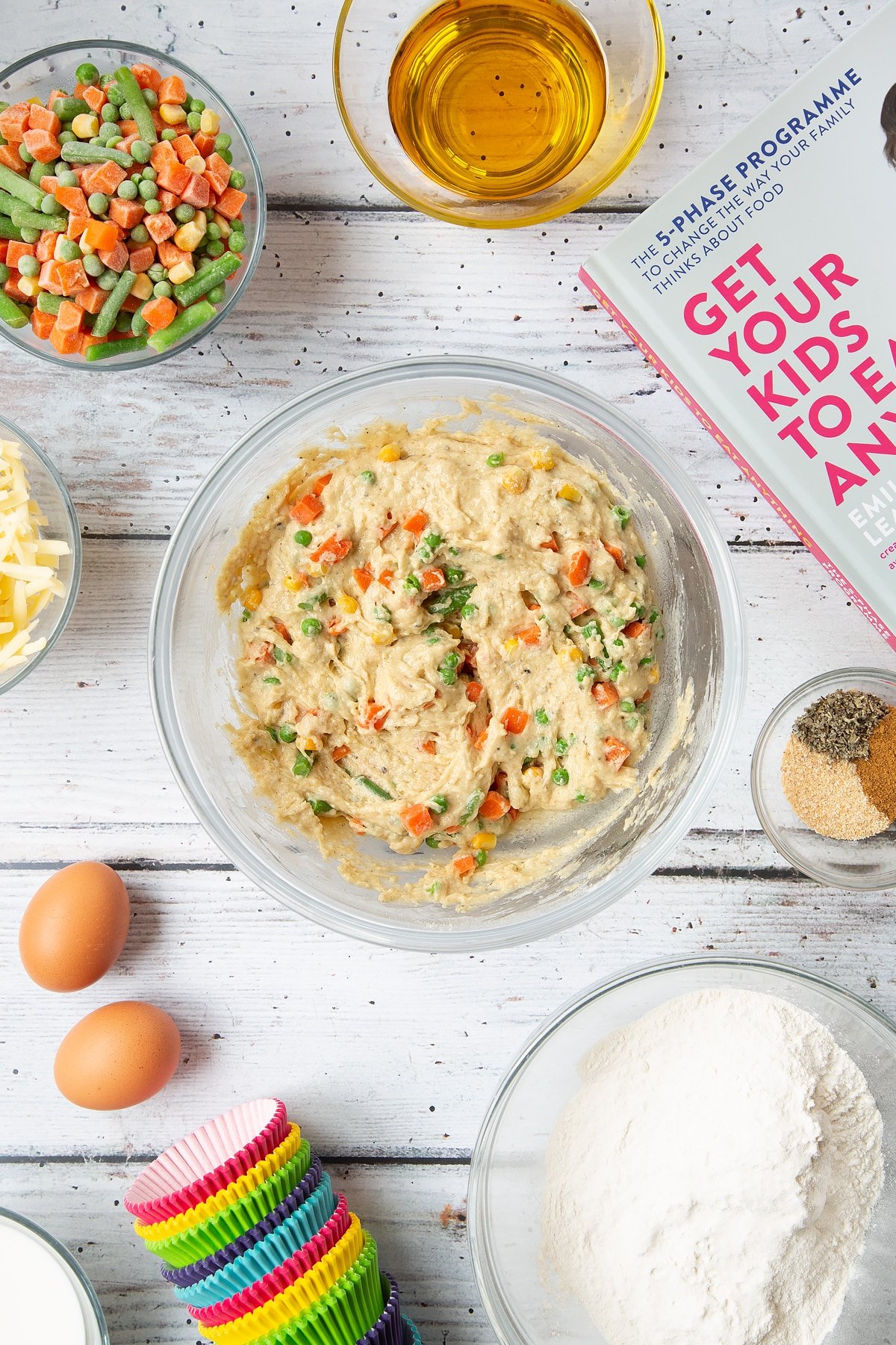 A mixing bowl containing a muffin batter studded with mixed chopped vegetables. The bowl is surrounded by the ingredients for savoury vegetable muffins.