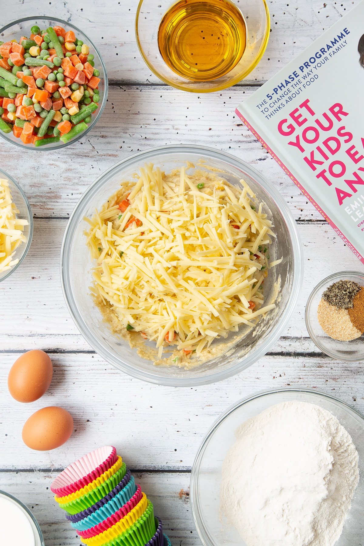 A mixing bowl containing a grated cheese on top of a muffin batter studded with mixed chopped vegetables. The bowl is surrounded by the ingredients for savoury vegetable muffins.