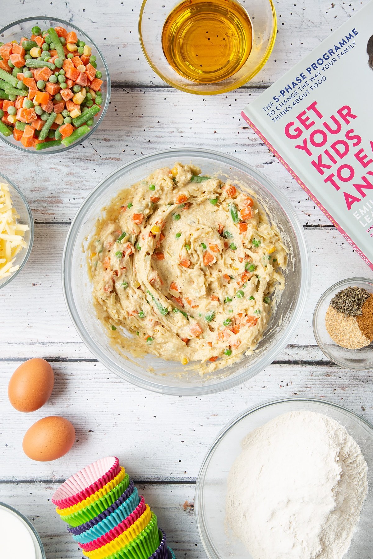 A mixing bowl containing a muffin batter with cheese and mixed chopped vegetables. The bowl is surrounded by the ingredients for savoury vegetable muffins.