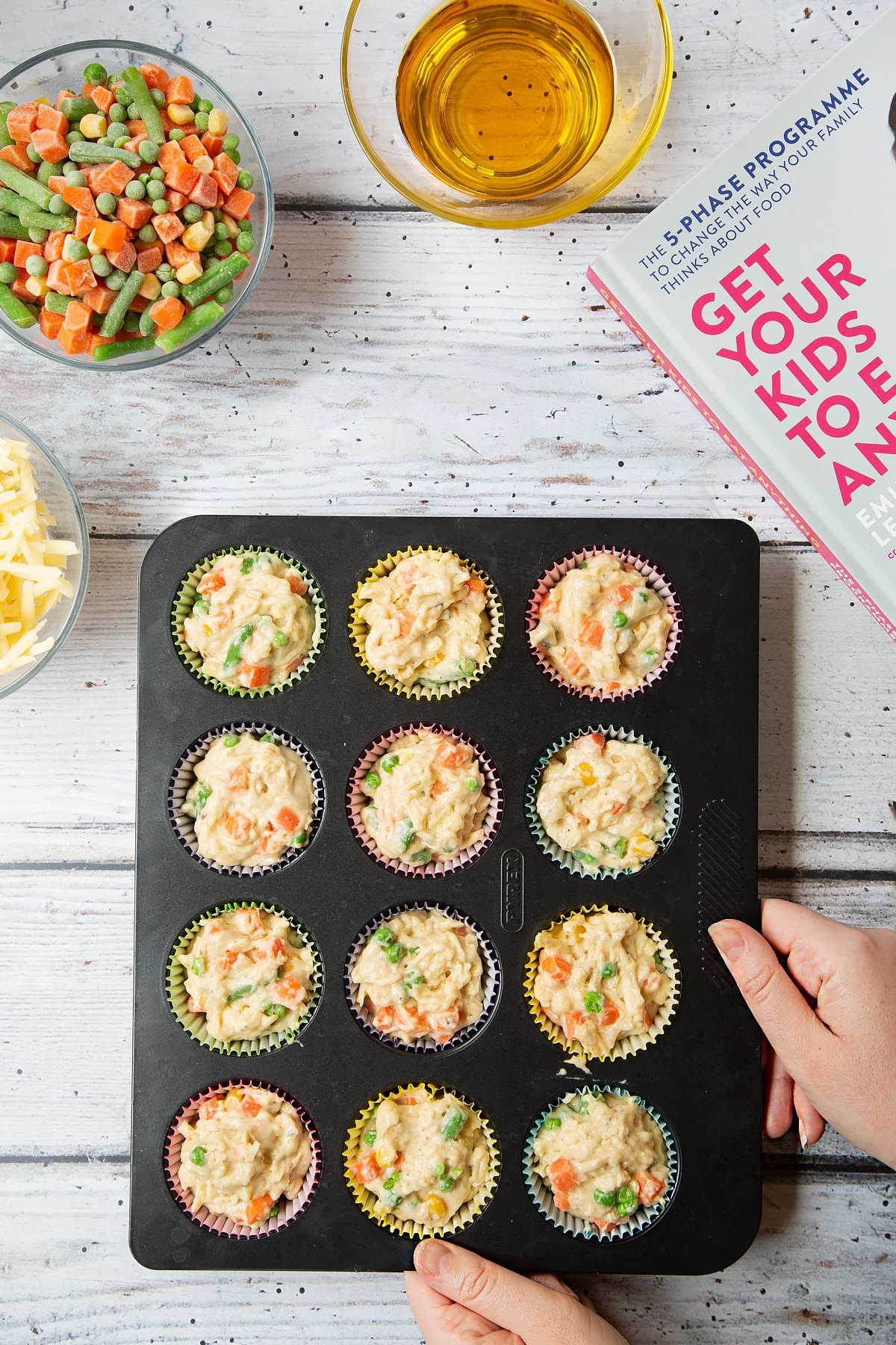 A 12-hole muffin tray lined with muffins cases and filled with savoury muffin batter with cheese and mixed chopped vegetables. The tray is surrounded by the ingredients for savoury vegetable muffins.