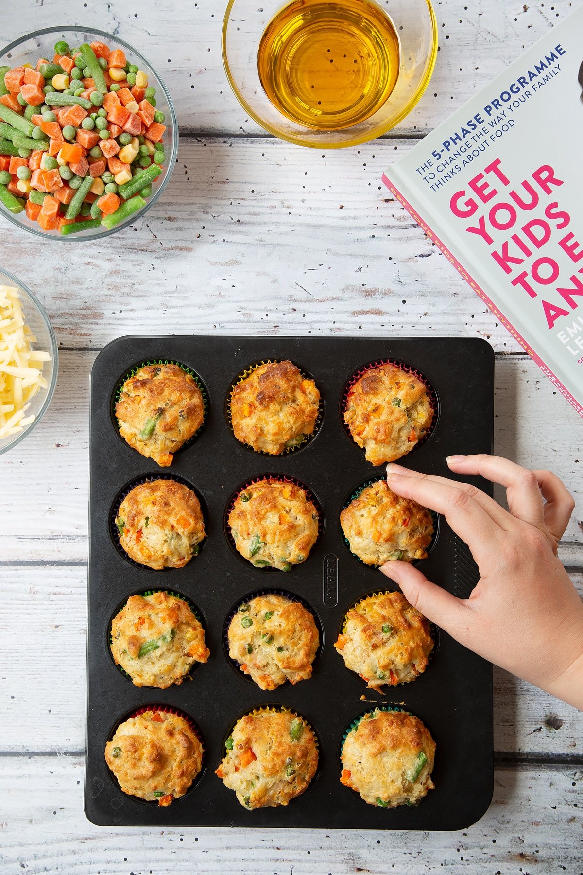 A 12-hole muffin tray filled with freshly baked savoury vegetable muffins, golden brown on top and studded with chopped vegetables. A hand reaches in to take a muffin.