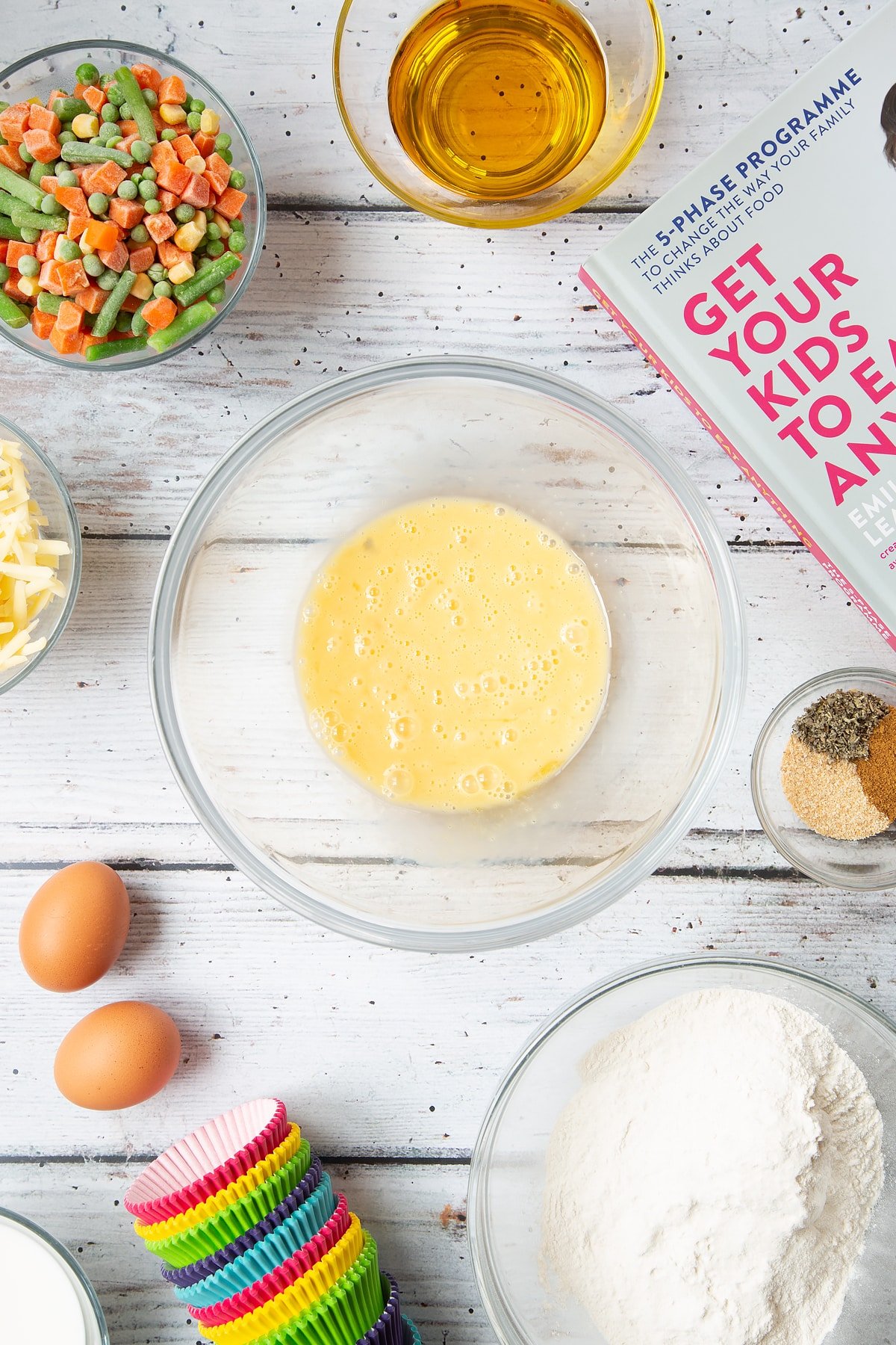 A mixing bowl containing beaten eggs. The bowl is surrounded by the ingredients for savoury vegetable muffins.