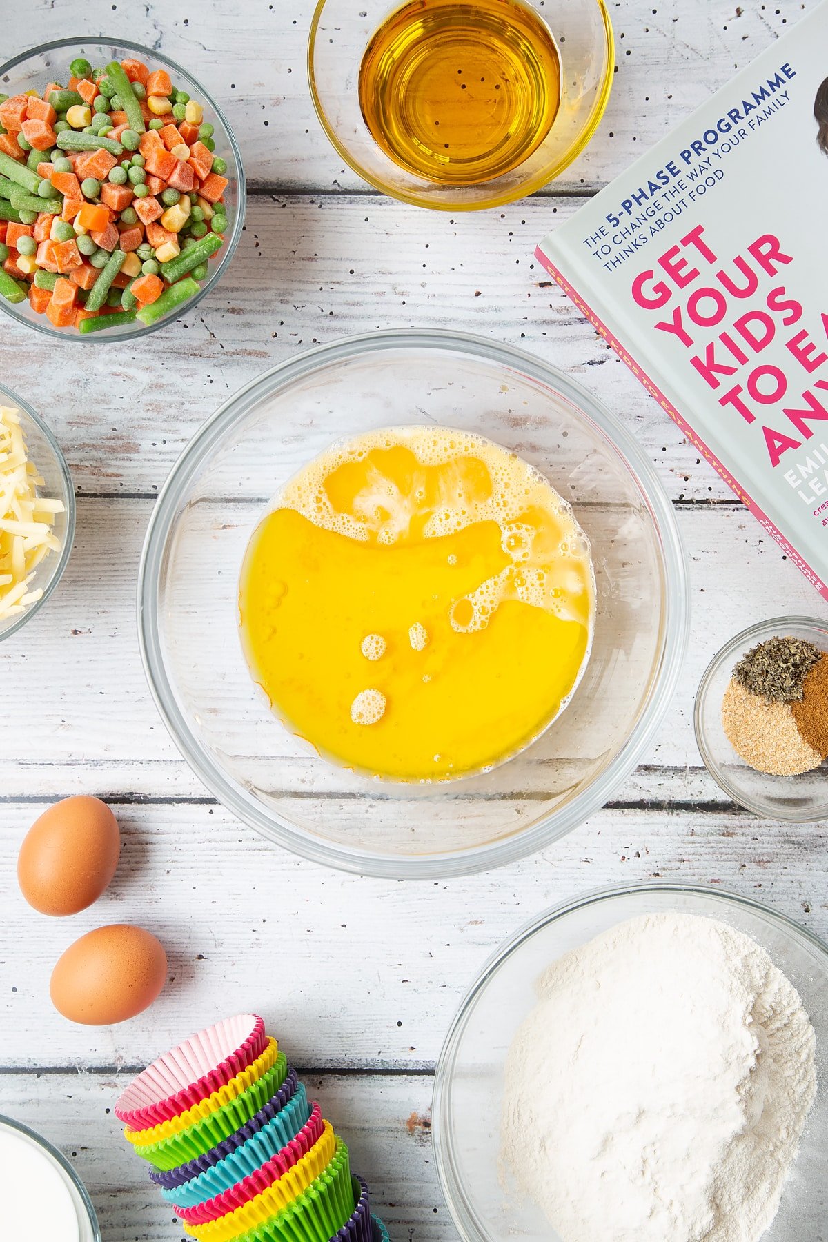 A mixing bowl containing beaten eggs, milk and oil. The bowl is surrounded by the ingredients for savoury vegetable muffins.