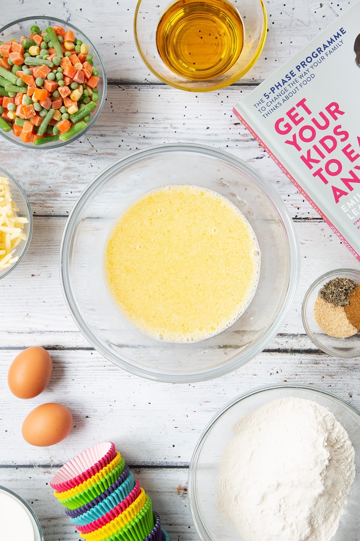 A mixing bowl containing eggs, milk and oil, beaten together. The bowl is surrounded by the ingredients for savoury vegetable muffins.