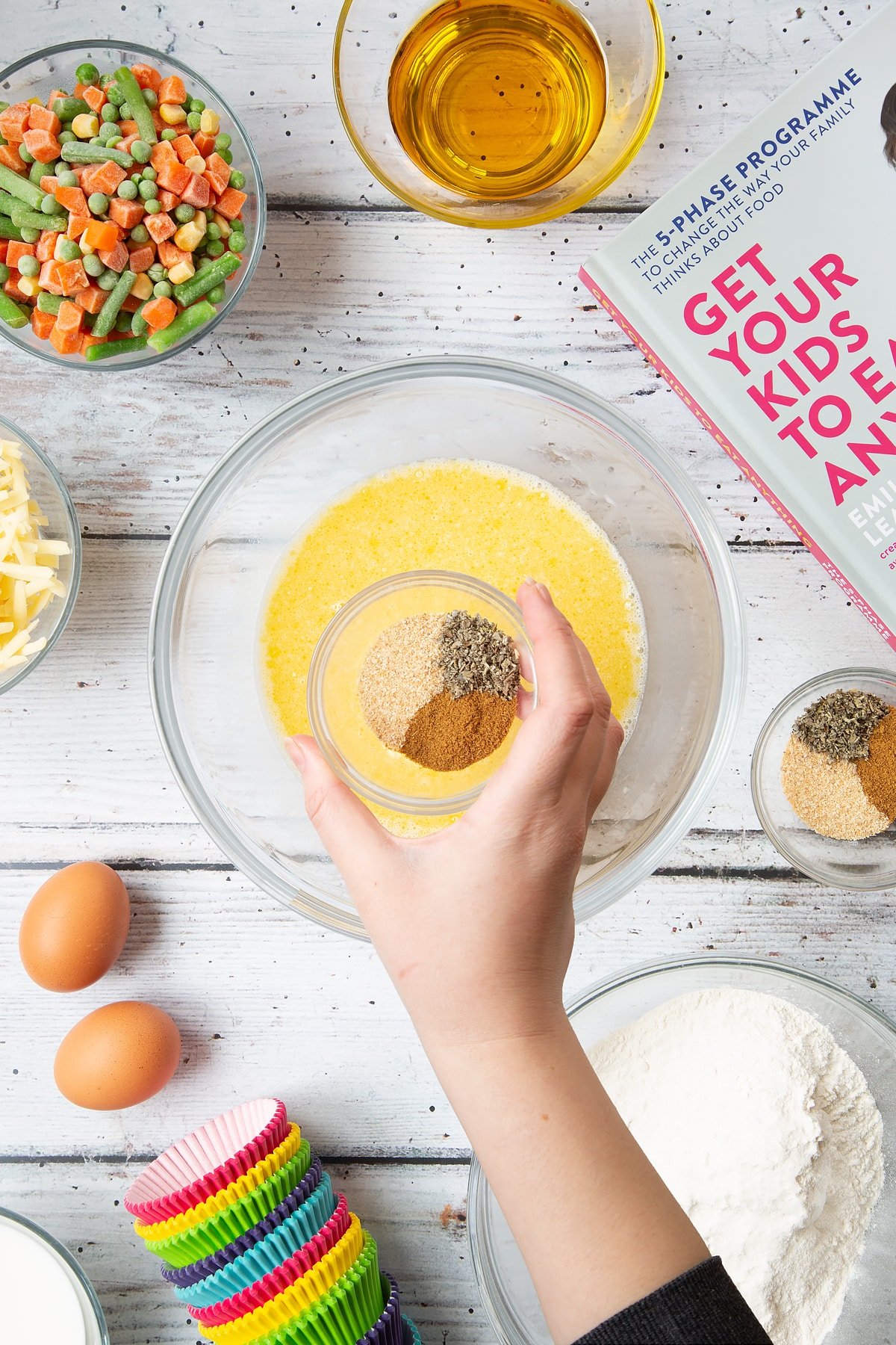 A mixing bowl containing eggs, milk and oil, beaten together. Above the bowl, a hand holds a small bowl containing garlic granules, dried sage and black pepper. The bowl is surrounded by the ingredients for savoury vegetable muffins.