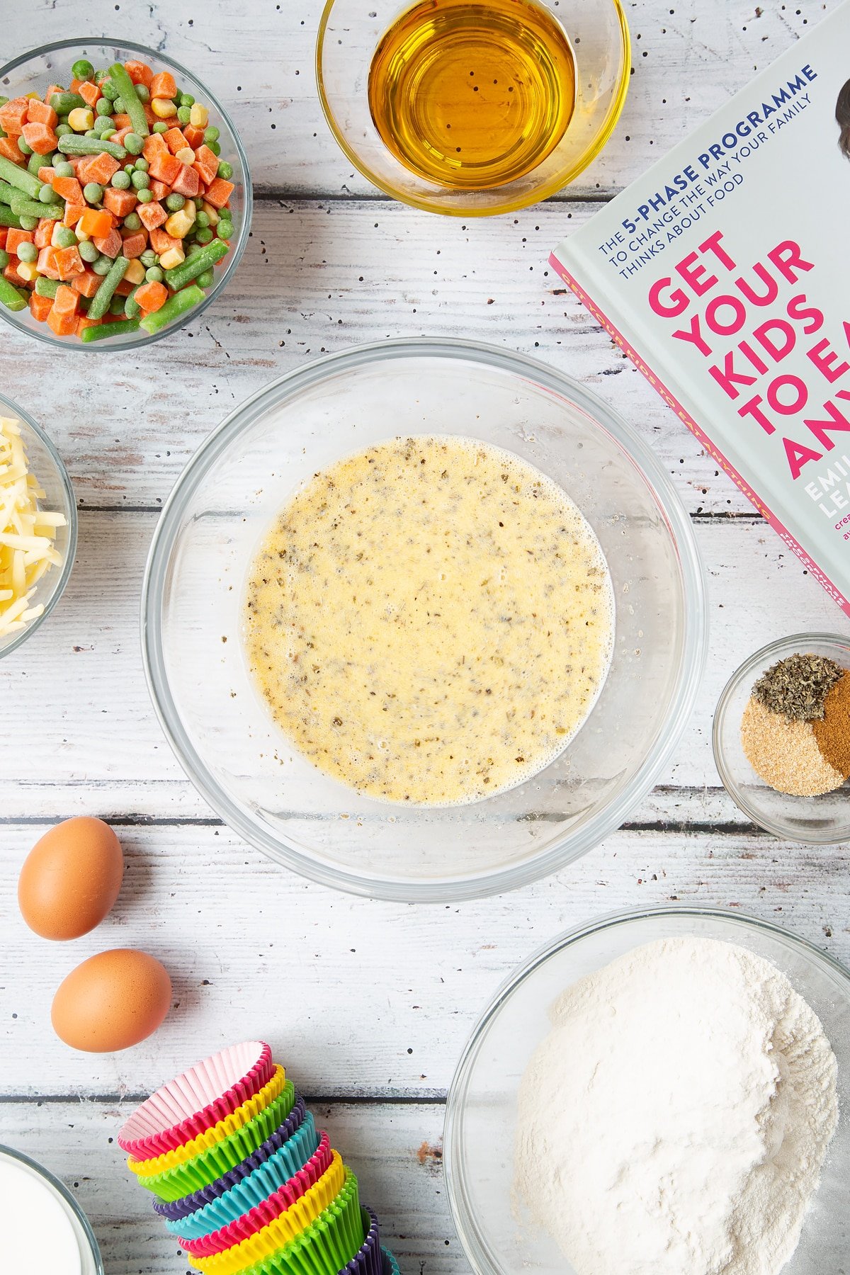 A mixing bowl containing eggs, milk and oil, garlic granules, dried sage and black pepper. The bowl is surrounded by the ingredients for savoury vegetable muffins.