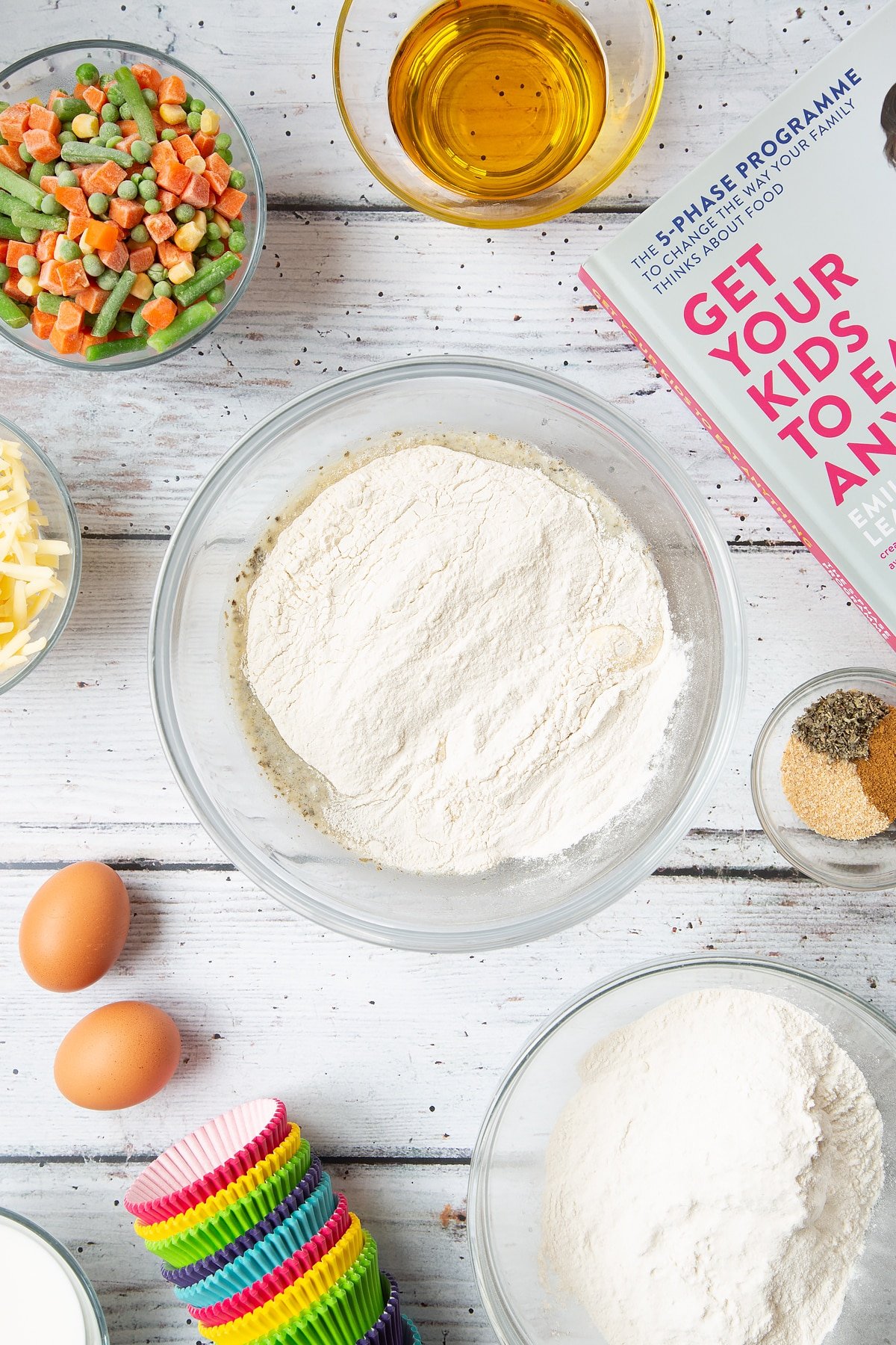 A mixing bowl containing flour on top of eggs, milk, oil, garlic granules, dried sage and black pepper. The bowl is surrounded by the ingredients for savoury vegetable muffins.