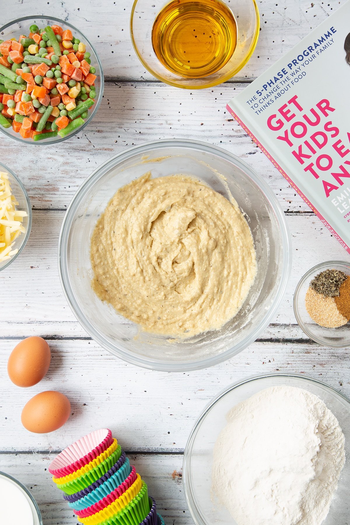 A mixing bowl containing a muffin batter made from flour, eggs, milk, oil, garlic granules, dried sage and black pepper. The bowl is surrounded by the ingredients for savoury vegetable muffins.