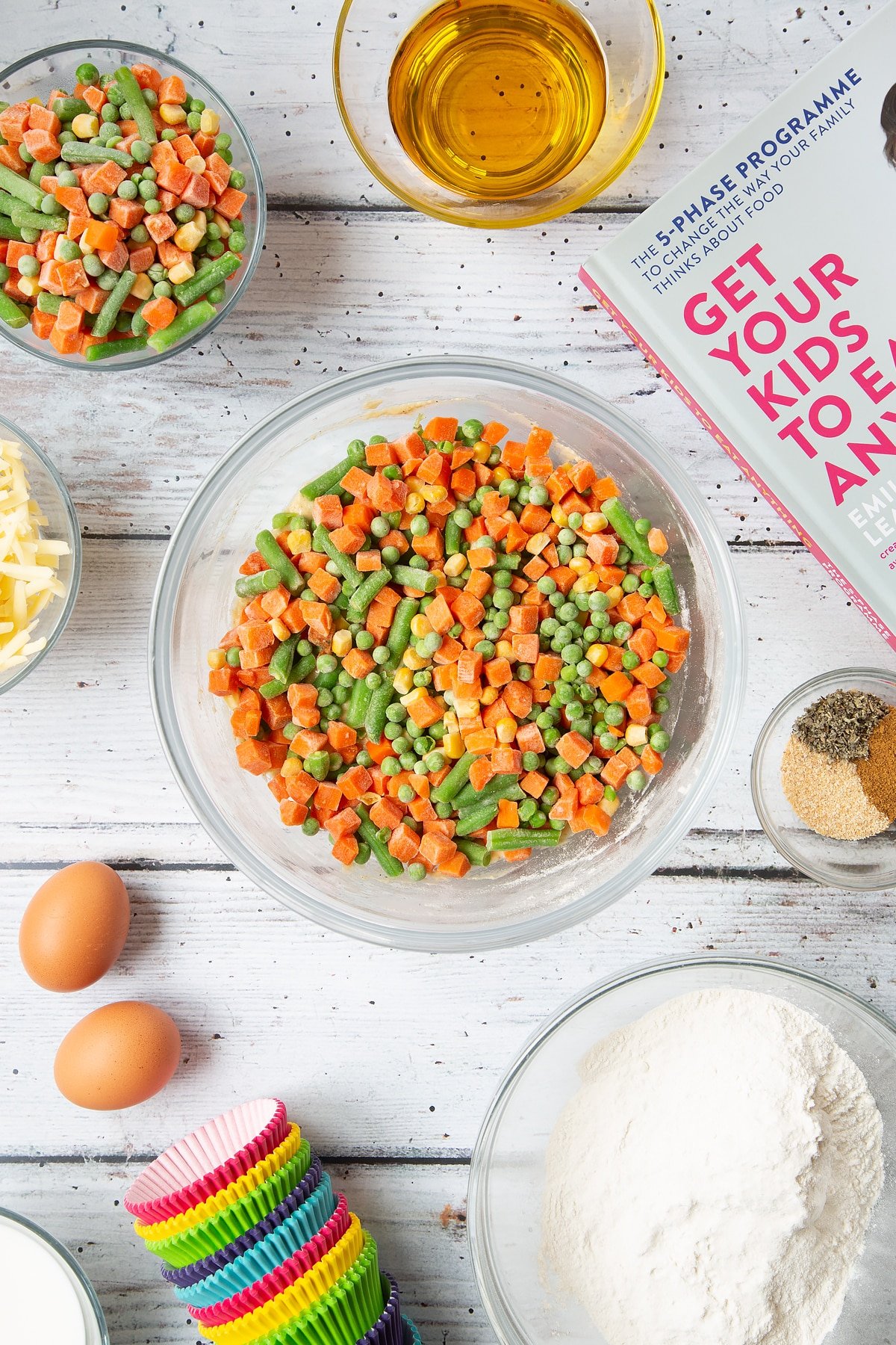 A mixing bowl containing a muffin batter topped with mixed chopped vegetables. The bowl is surrounded by the ingredients for savoury vegetable muffins.