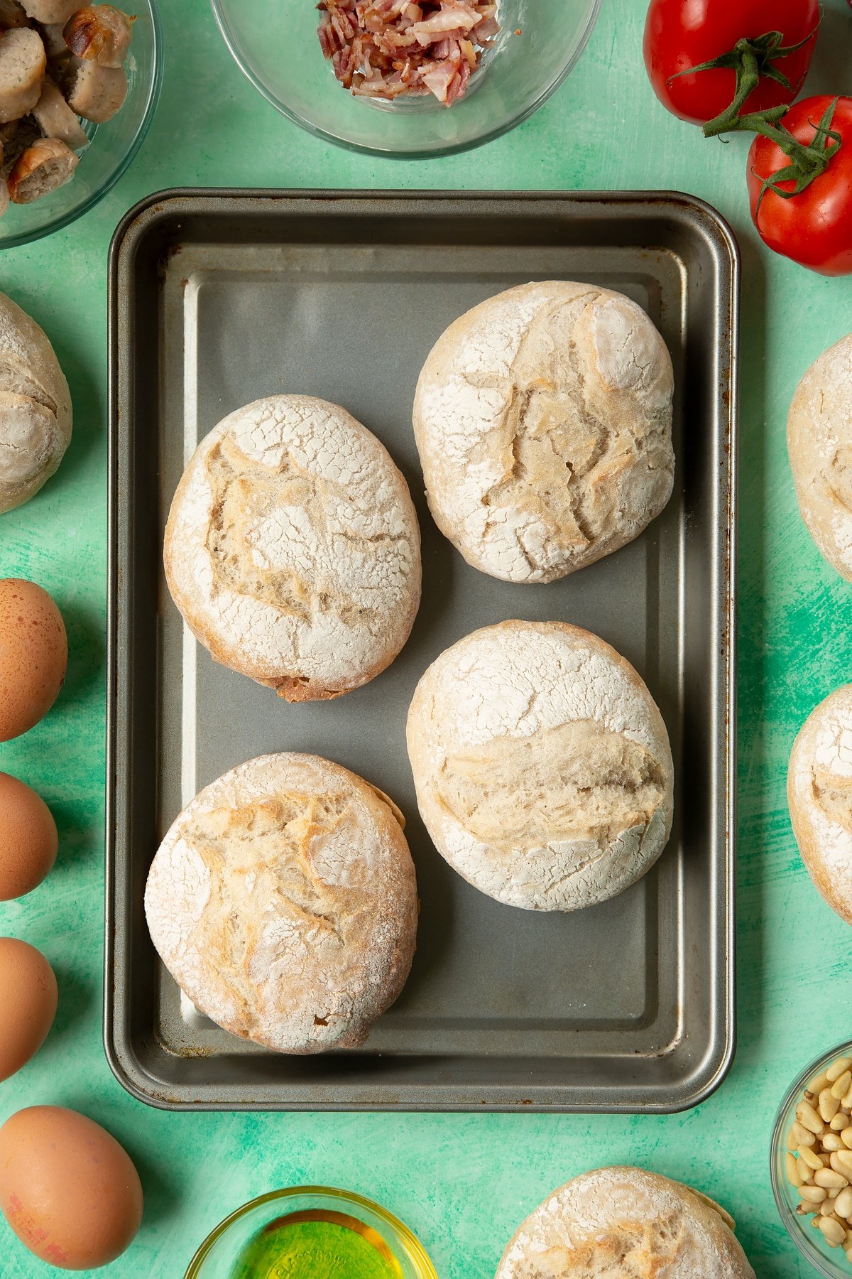 Four crusty bread rolls on a baking tray. The tray is surrounded by ingredients to make breakfast rolls.