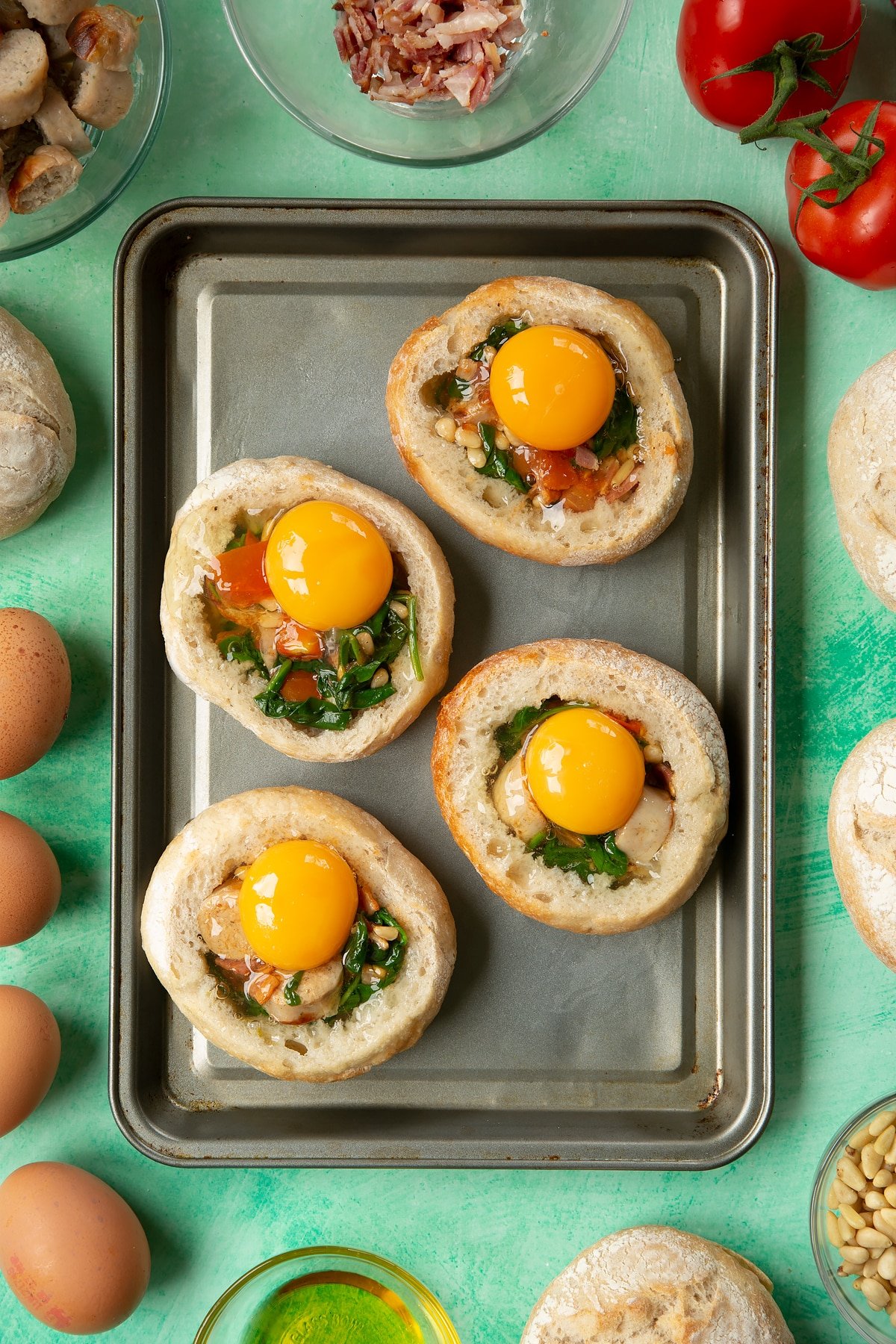 Four bread rolls on a baking tray. The rolls are hollowed out and filled with a mix of fried sausage, bacon, chopped tomatoes, pine nuts and wilted spinach, and topped with eggs. The tray is surrounded by ingredients to make breakfast rolls.