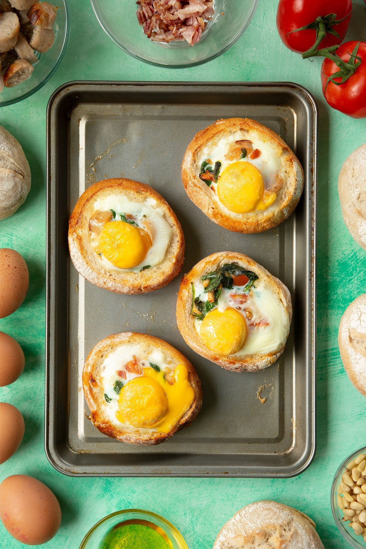 Four bread rolls on a baking tray. The rolls are hollowed out and filled with a mix of fried sausage, bacon, chopped tomatoes, pine nuts and wilted spinach, and topped with baked eggs. The tray is surrounded by ingredients to make breakfast rolls.