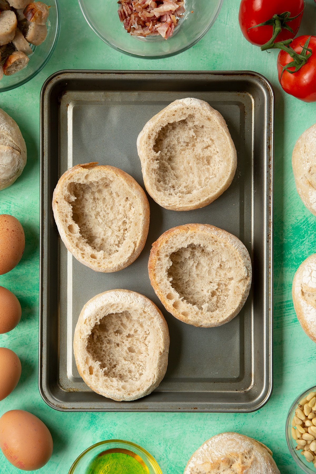 Four crusty bread rolls on a baking tray. The tops have been cut off and most of the inner crumb removed. The tray is surrounded by ingredients to make breakfast rolls.