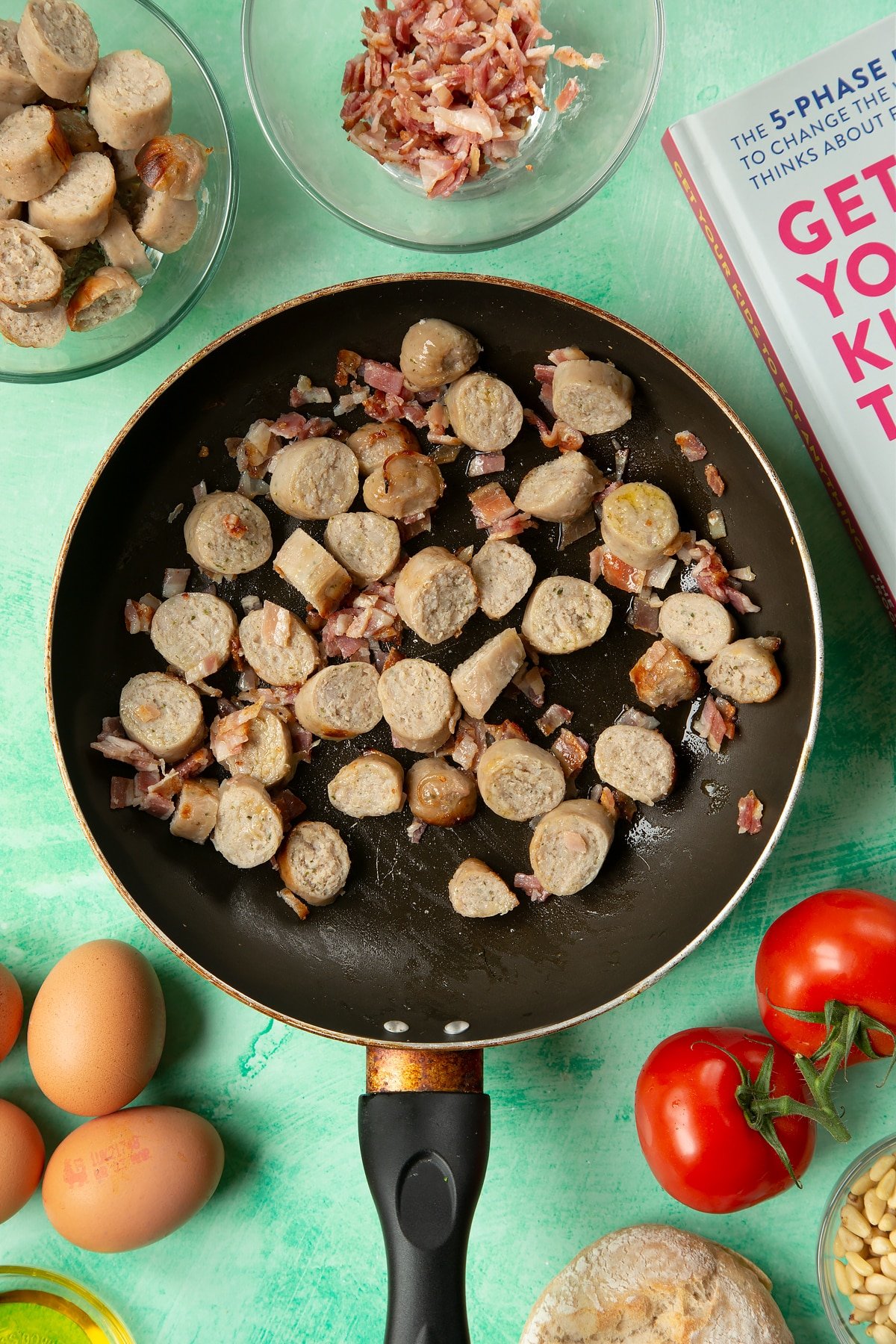 A frying pan containing chopped sausage and bacon. The pan is surrounded by ingredients to make breakfast rolls.