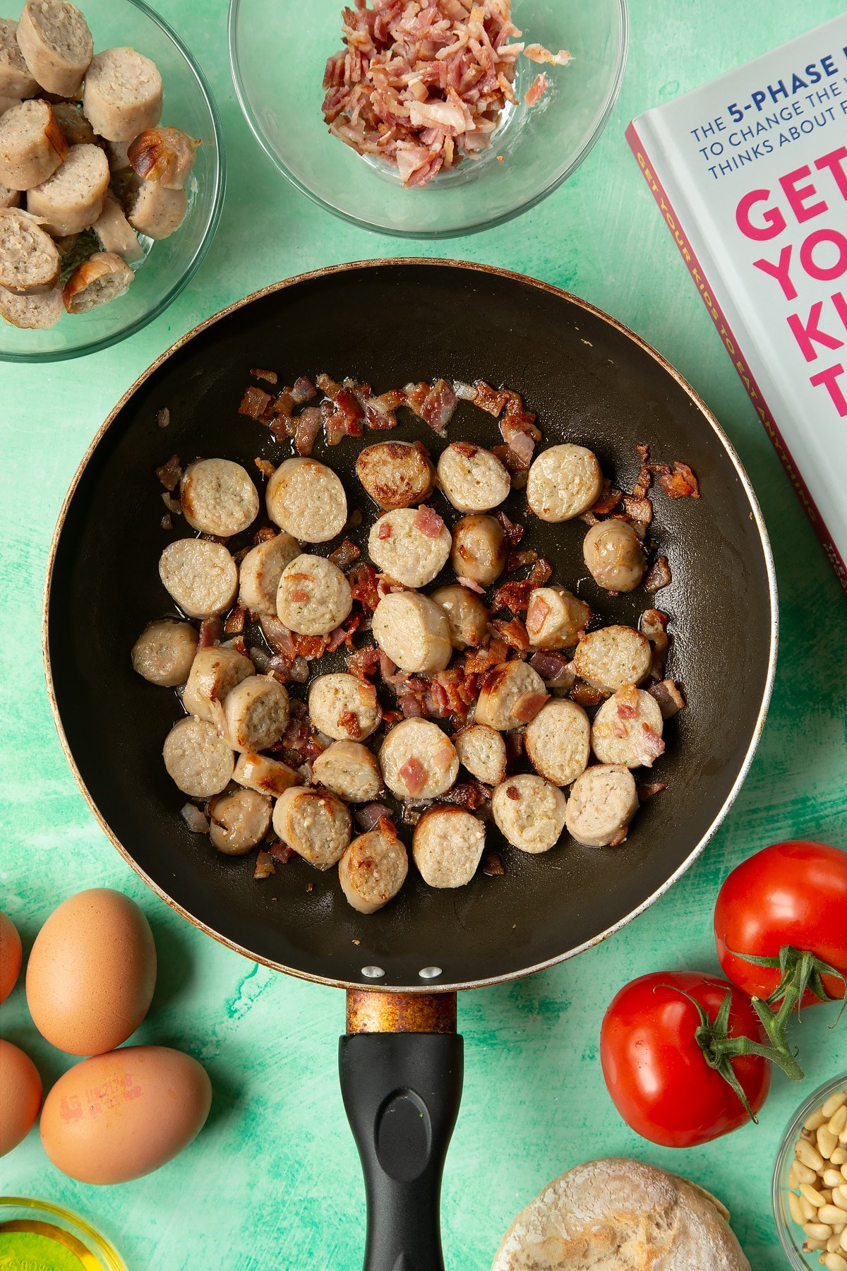 A frying pan containing fried sausage and bacon. The pan is surrounded by ingredients to make breakfast rolls.