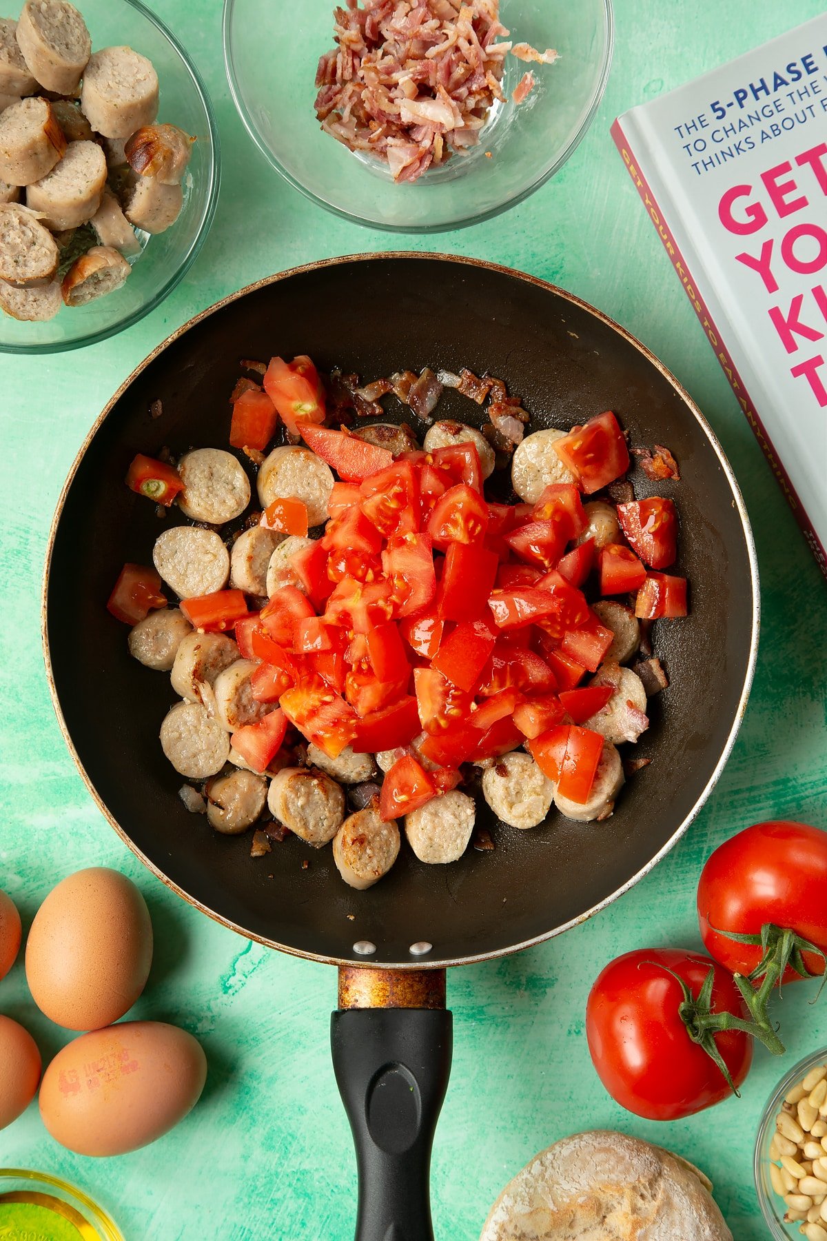 A frying pan containing fried sausage and bacon, topped with chopped tomatoes. The pan is surrounded by ingredients to make breakfast rolls.