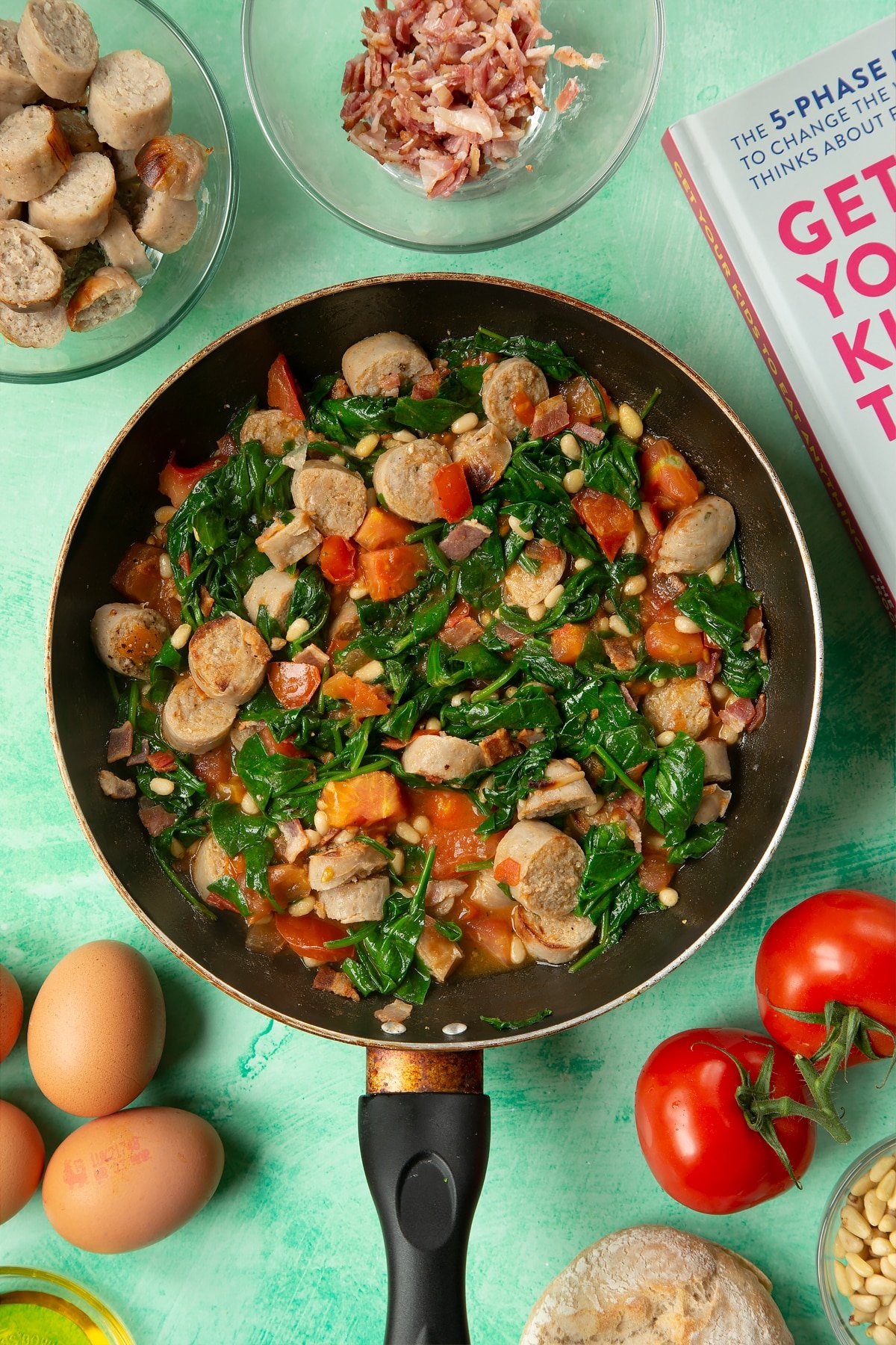 A frying pan containing fried sausage, bacon, chopped tomatoes, pine nuts and wilted spinach. The pan is surrounded by ingredients to make breakfast rolls.