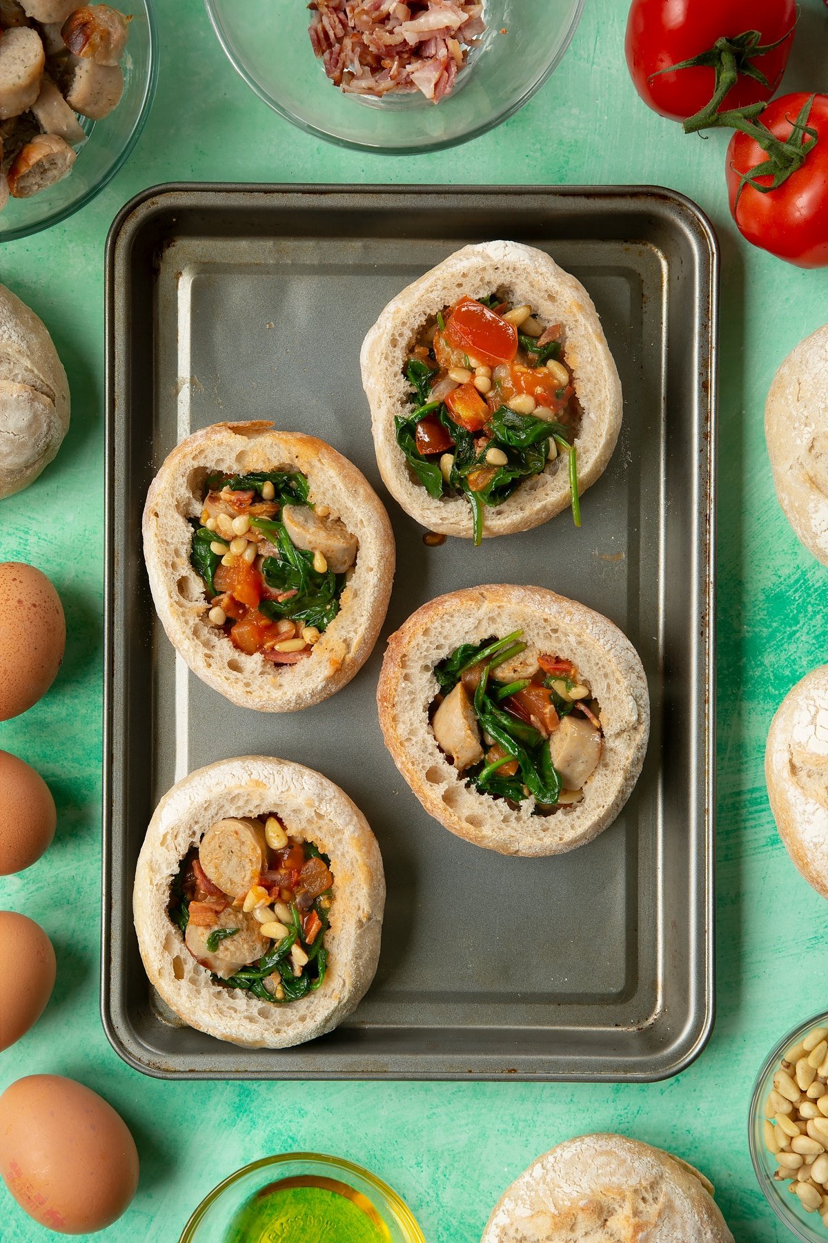 Four bread rolls on a baking tray. The rolls are hollowed out and filled with a mix of fried sausage, bacon, chopped tomatoes, pine nuts and wilted spinach. The tray is surrounded by ingredients to make breakfast rolls.