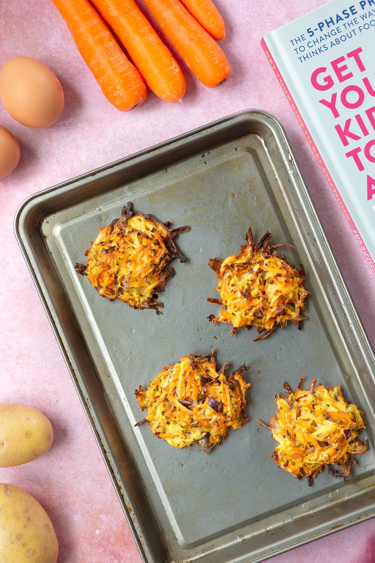 Four cooked carrot patties on an oiled baking tray. The tray is surrounded by ingredients to make carrot patties.