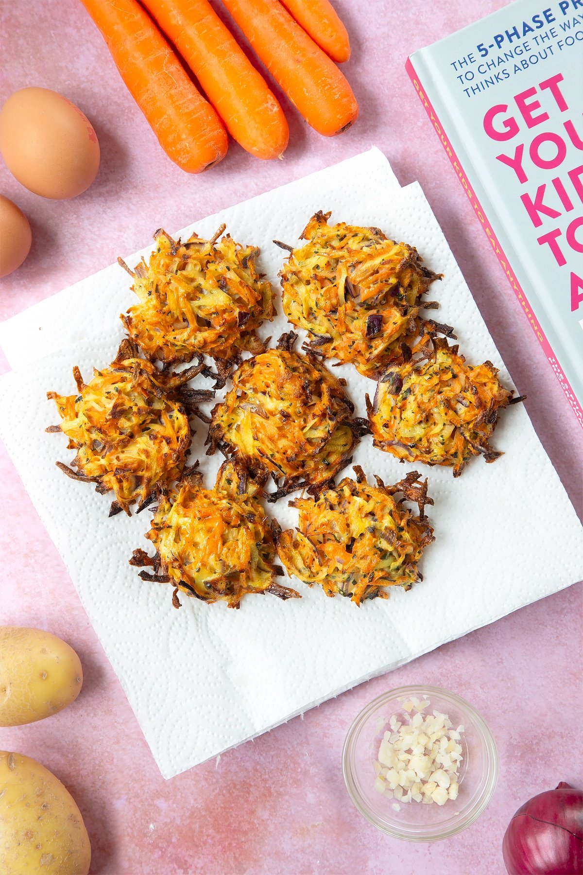 Seven cooked carrot patties on a plate lined with kitchen paper. The plate is surrounded by ingredients to make carrot patties.