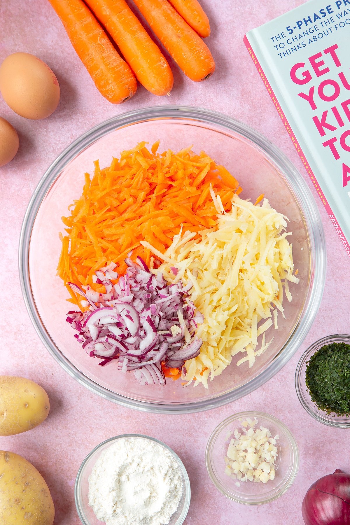 Grated carrot, red onion and potato in a mixing bowl. The bowl is surrounded by ingredients to make carrot patties.