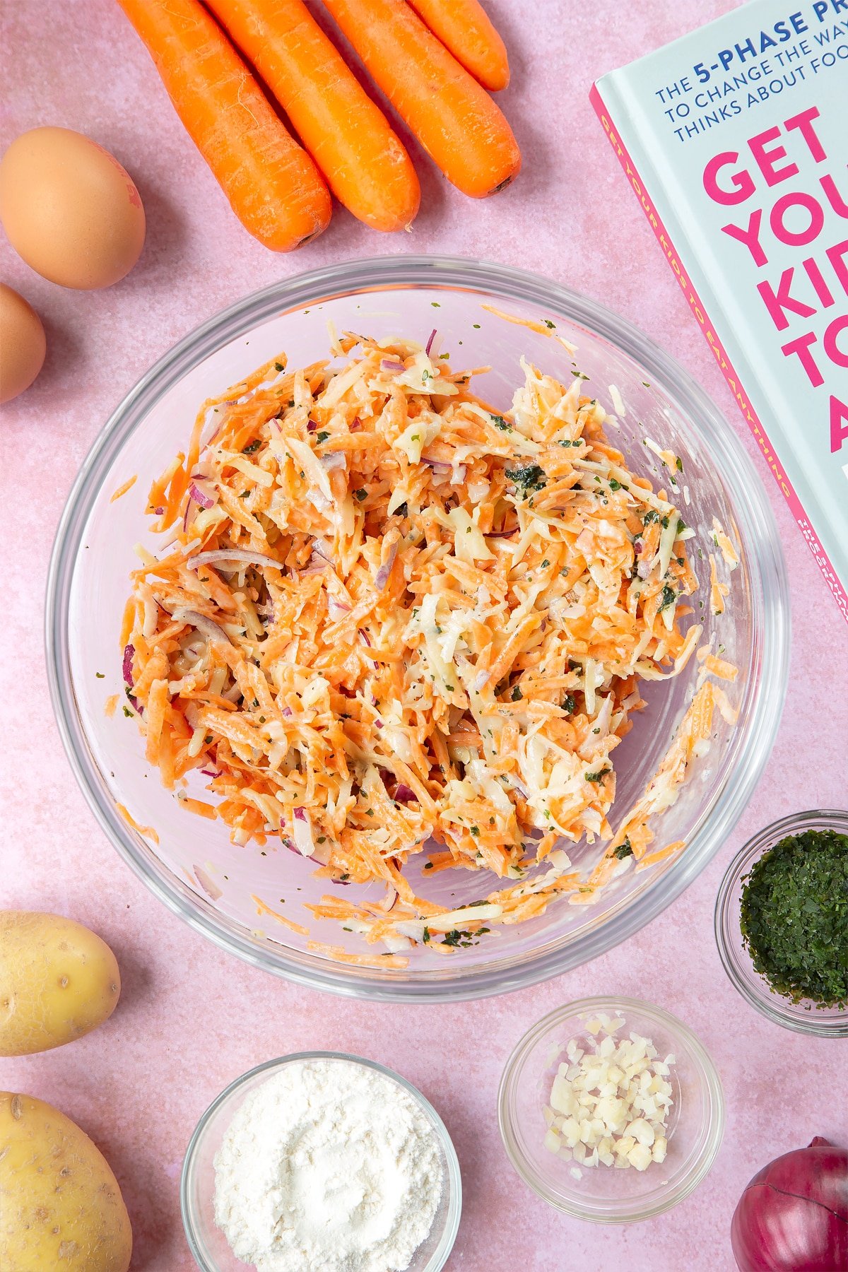 Grated carrot, potato, red onion and coriander in a flour and egg batter in a mixing bowl. The bowl is surrounded by ingredients to make carrot patties.