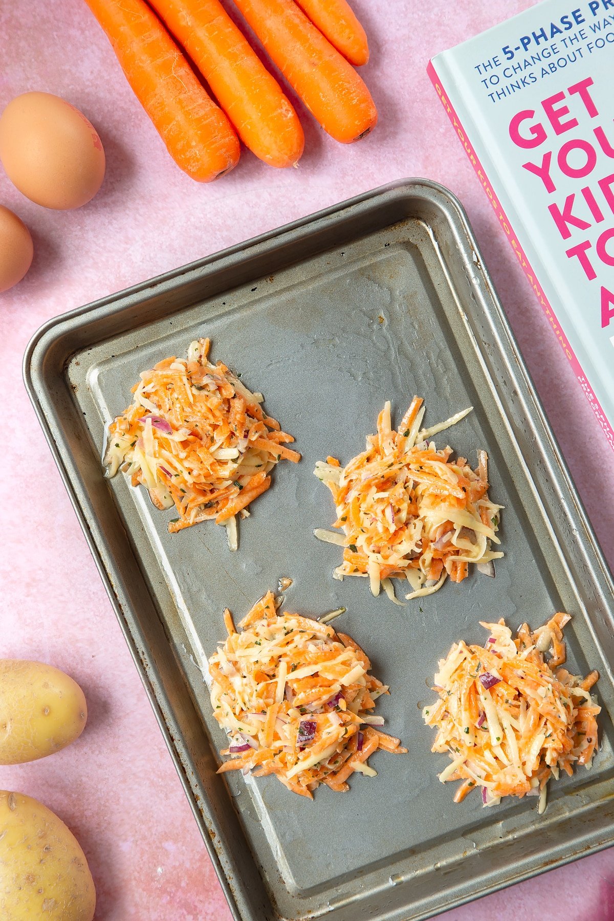 Four mounds of carrot patty mix on an oiled baking tray. The tray is surrounded by ingredients to make carrot patties.
