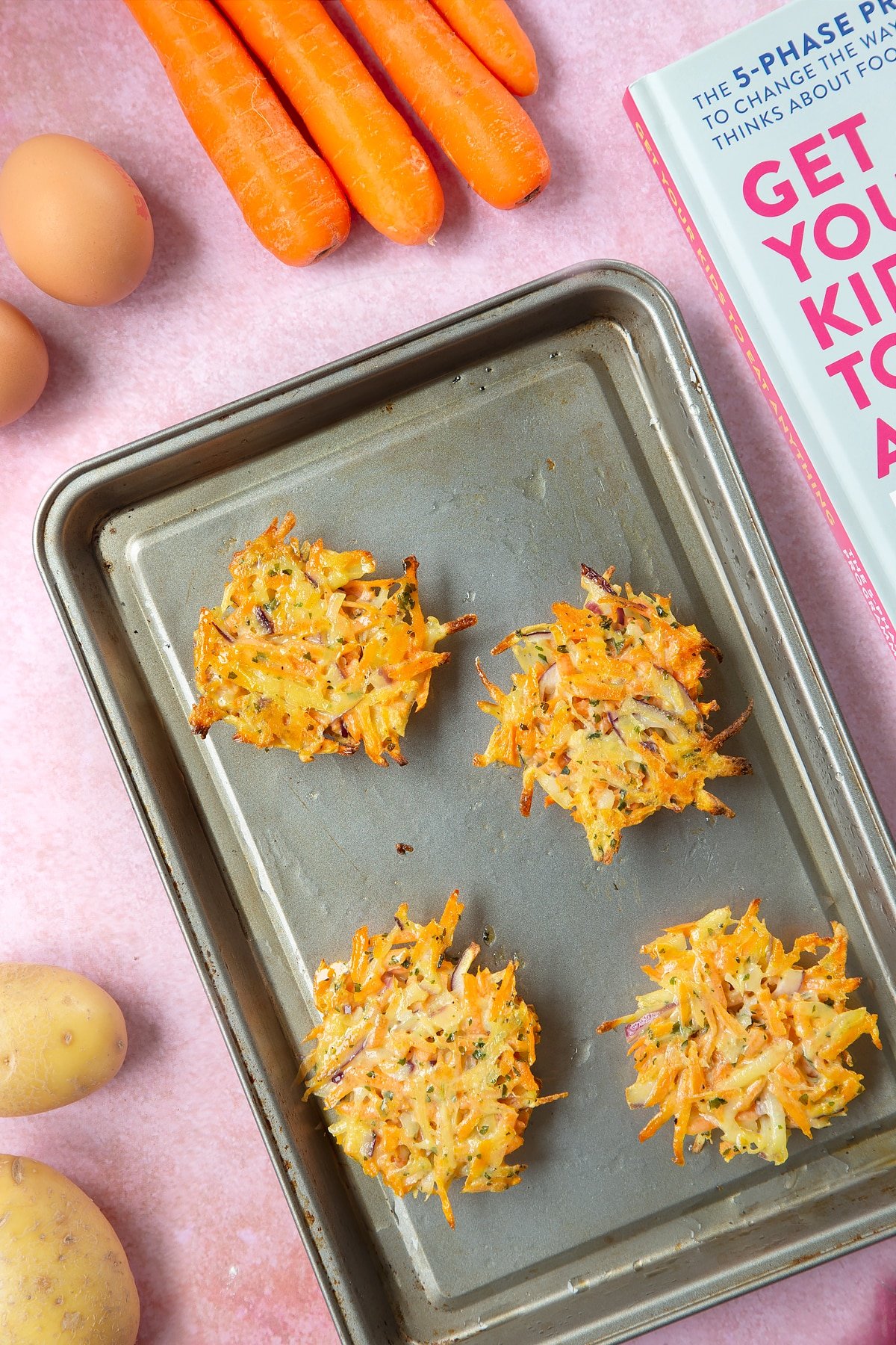 Four mounds of carrot patty mix on an oiled baking tray. The patties are flipped over to show the semi-cooked undersides. The tray is surrounded by ingredients to make carrot patties.