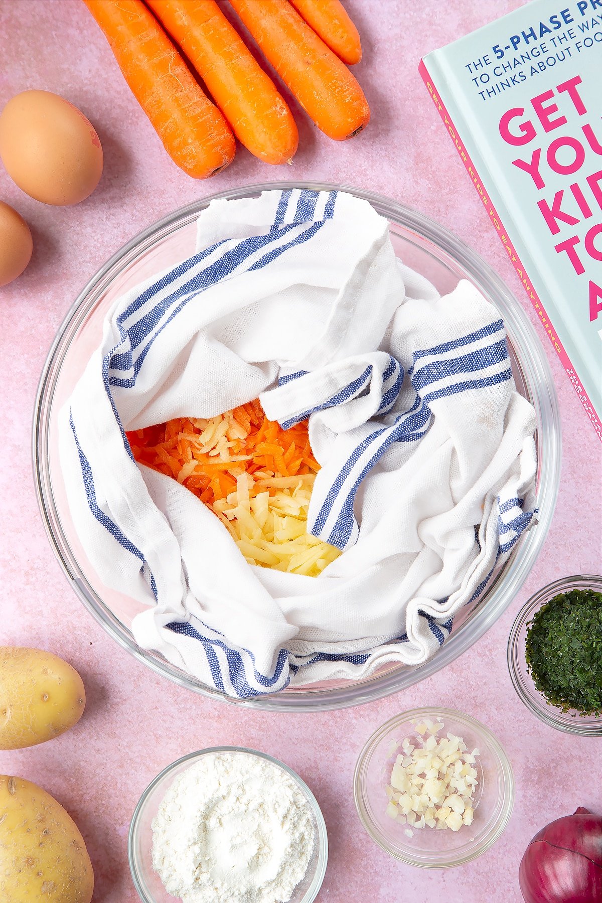 Grated carrot, red onion and potato inside a white and blue tea towel in a mixing bowl. The bowl is surrounded by ingredients to make carrot patties.