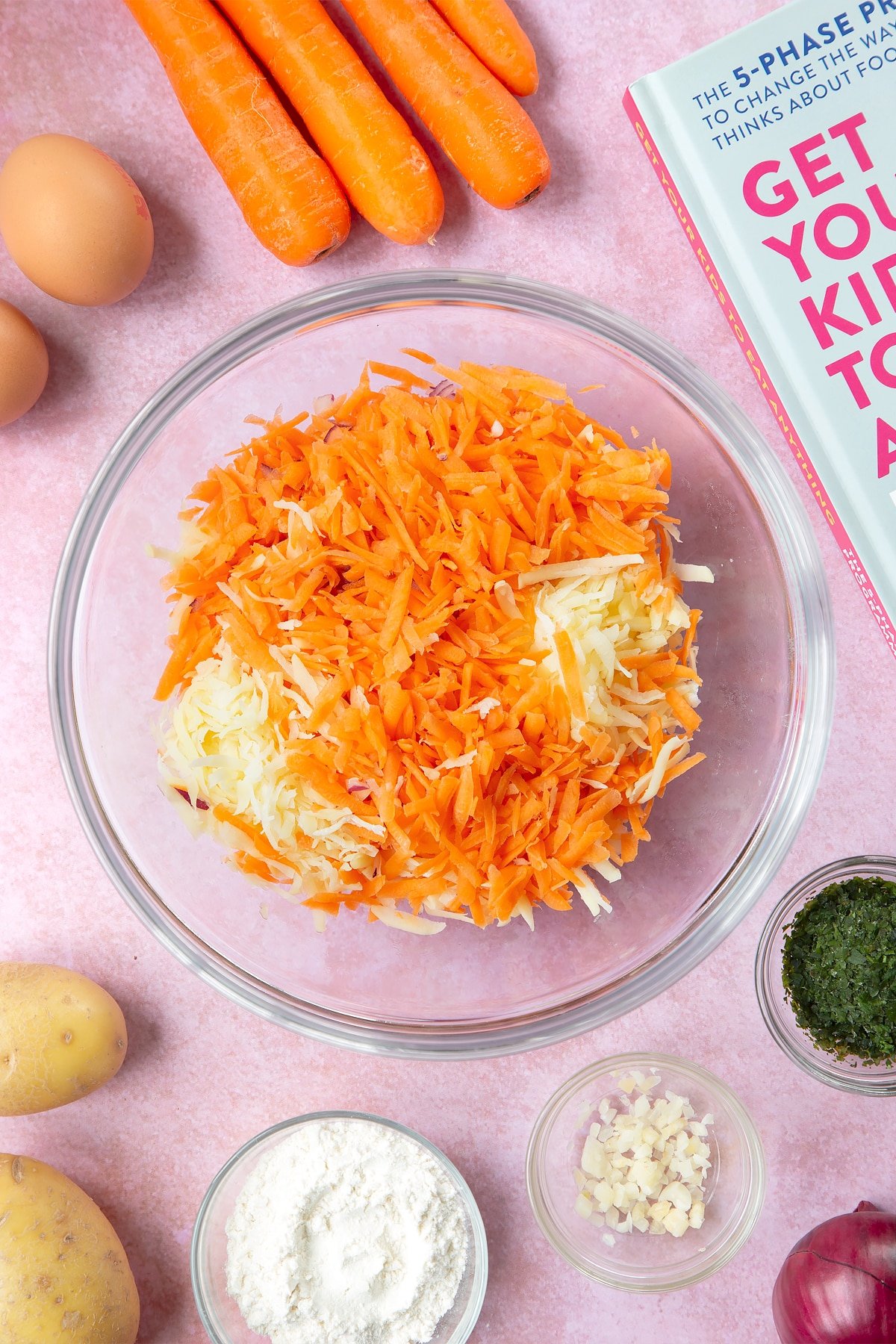 Grated carrot, red onion and potato in a mixing bowl. Excess moisture has been removed from the veggies. The bowl is surrounded by ingredients to make carrot patties.