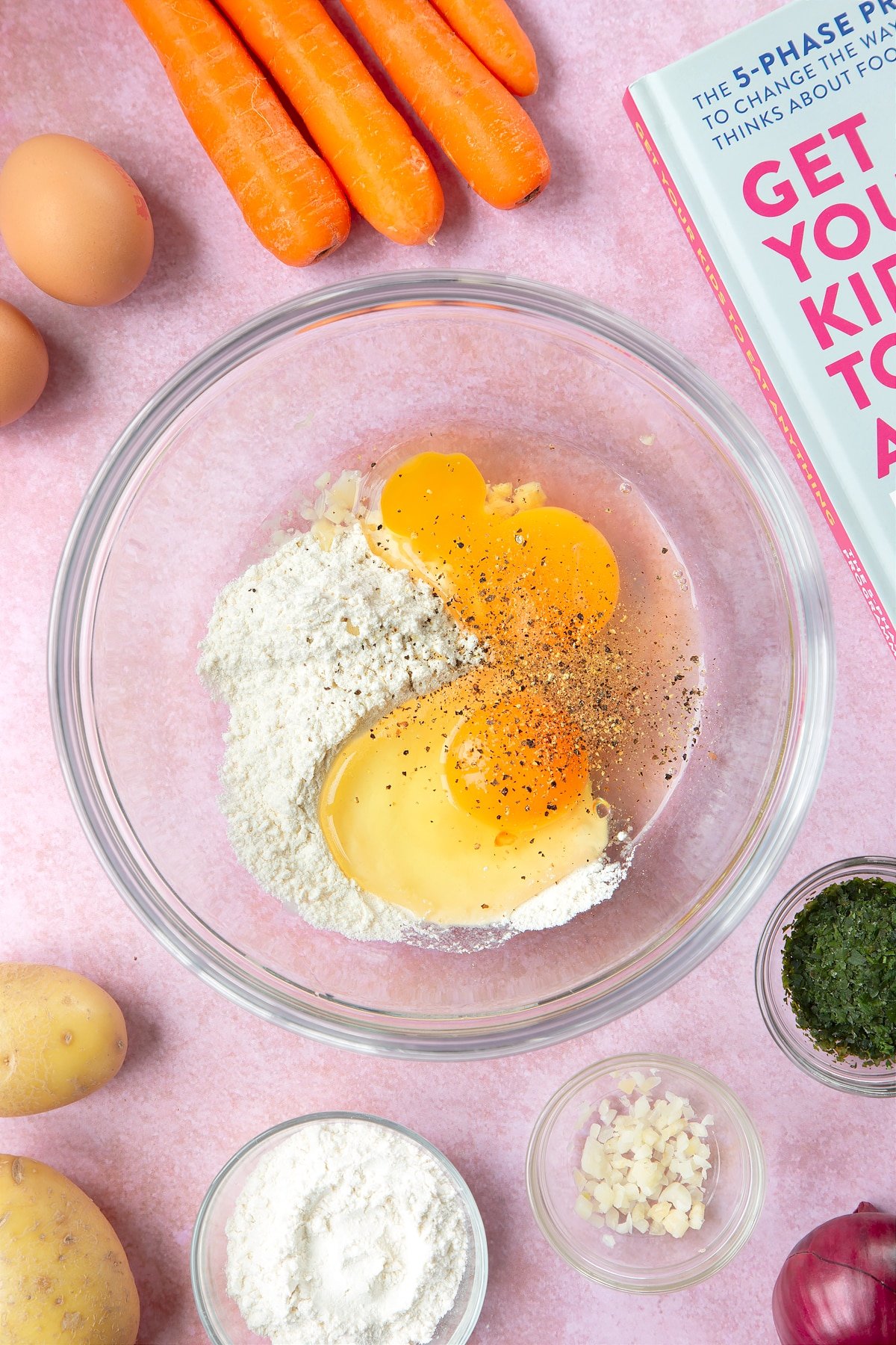 Flour, eggs, salt and pepper in a mixing bowl. The bowl is surrounded by ingredients to make carrot patties.