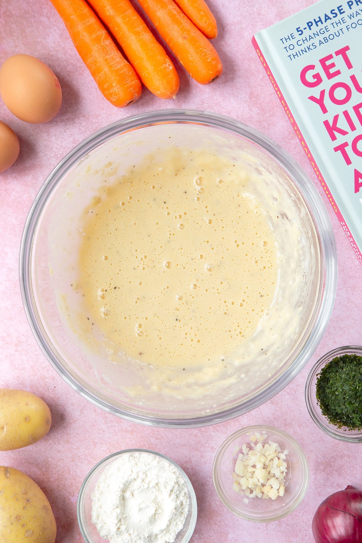 A batter made from flour, eggs, salt and pepper in a mixing bowl. The bowl is surrounded by ingredients to make carrot patties.