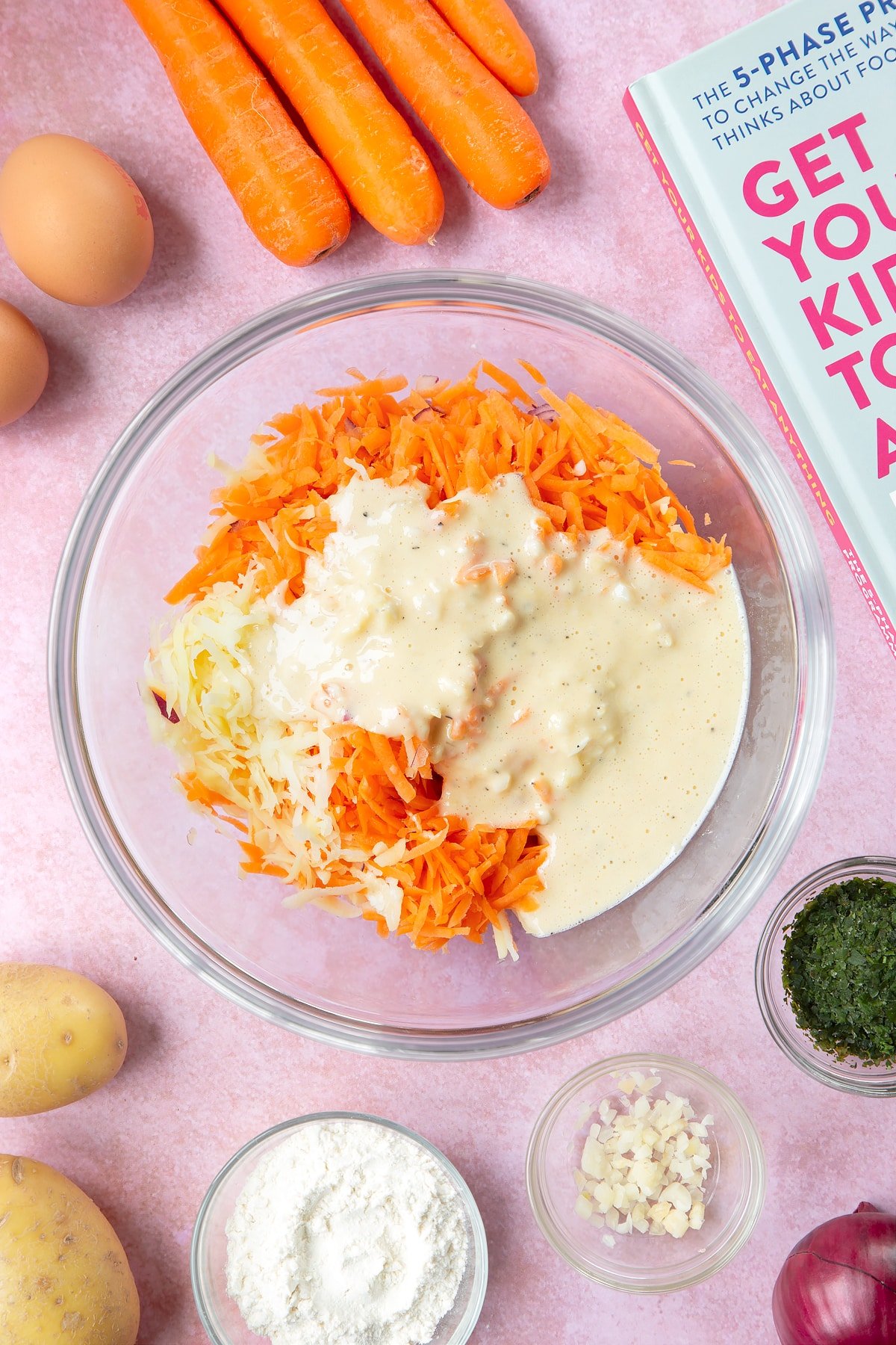 Grated carrot, potato and red onion topped with a batter made from flour, eggs, salt and pepper in a mixing bowl. The bowl is surrounded by ingredients to make carrot patties.