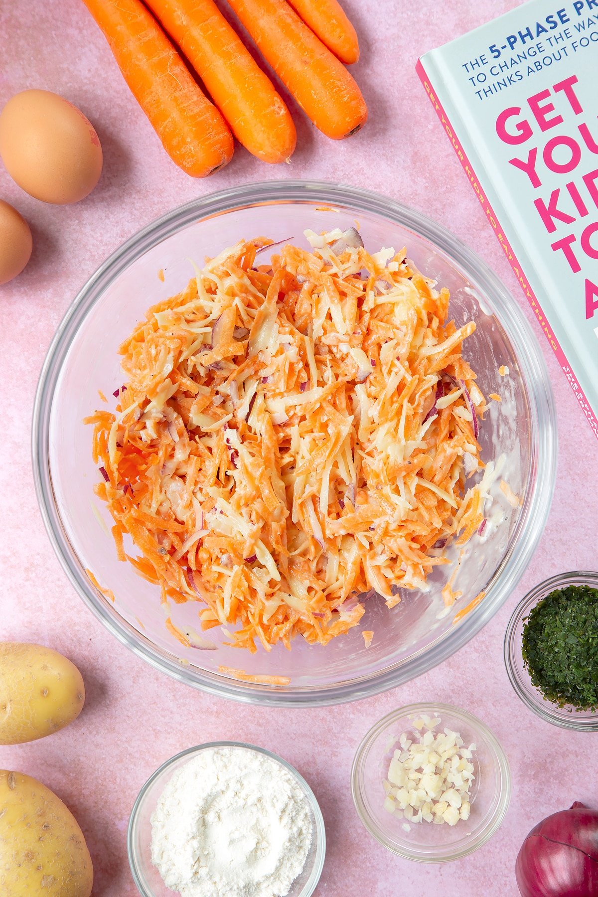 Grated carrot, potato and red onion mixed with a batter made from flour, eggs, salt and pepper in a mixing bowl. The bowl is surrounded by ingredients to make carrot patties.