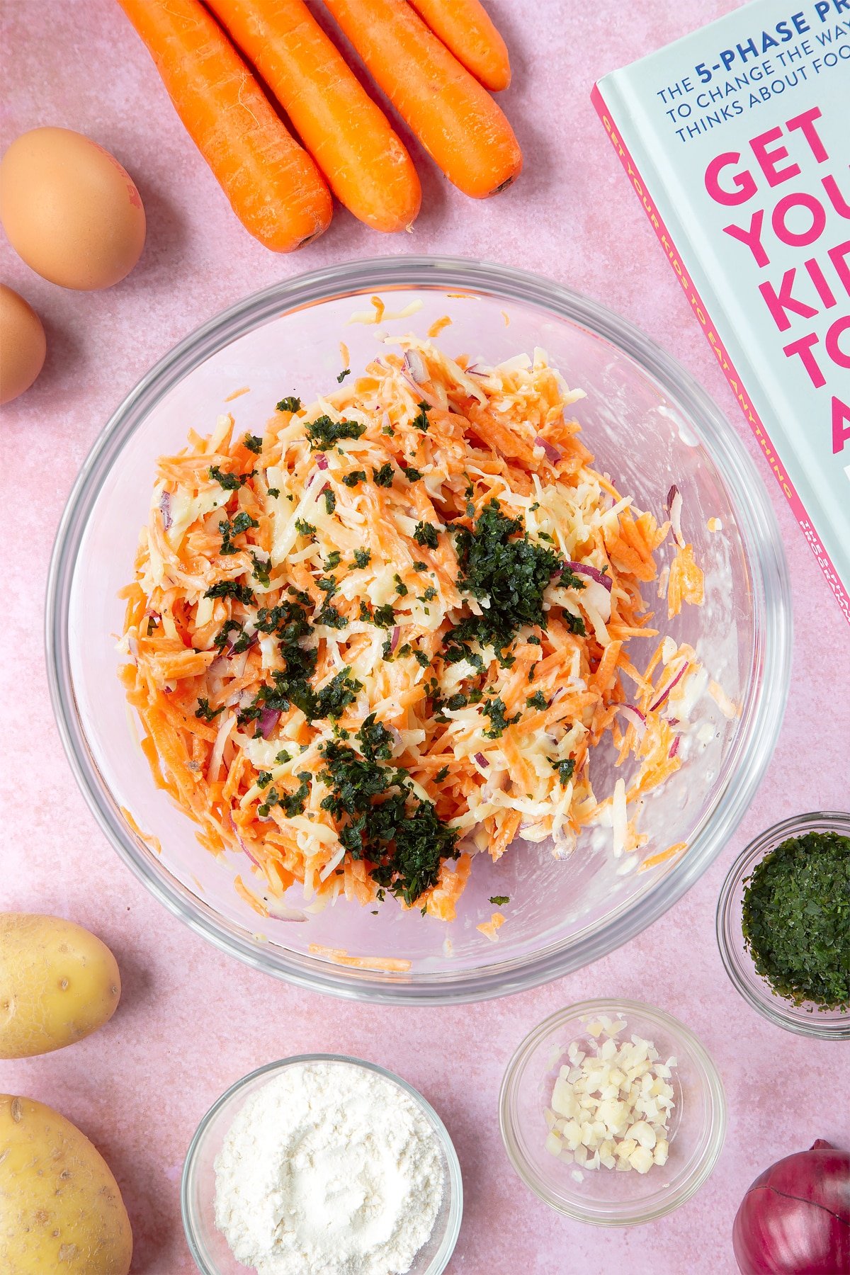 Grated carrot, potato and red onion in a batter, topped with chopped coriander in a mixing bowl. The bowl is surrounded by ingredients to make carrot patties.