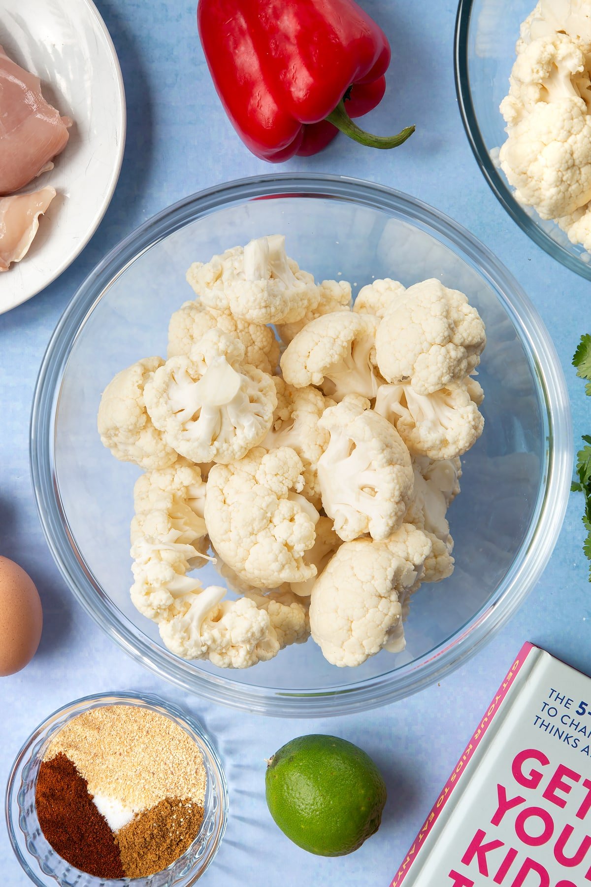 A glass mixing bowl filled with cauliflower florets. The bowl is surrounded by ingredients for cauliflower tacos. 