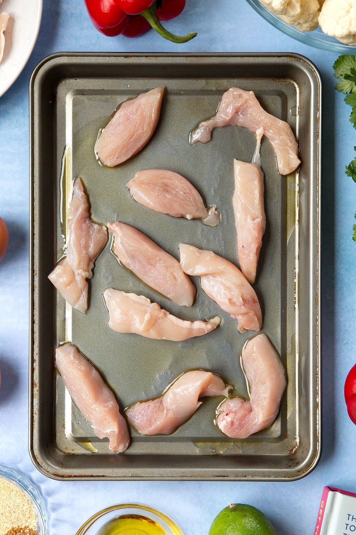 Sliced chicken breast on an oiled tray. The tray is surrounded by ingredients for cauliflower tacos. 